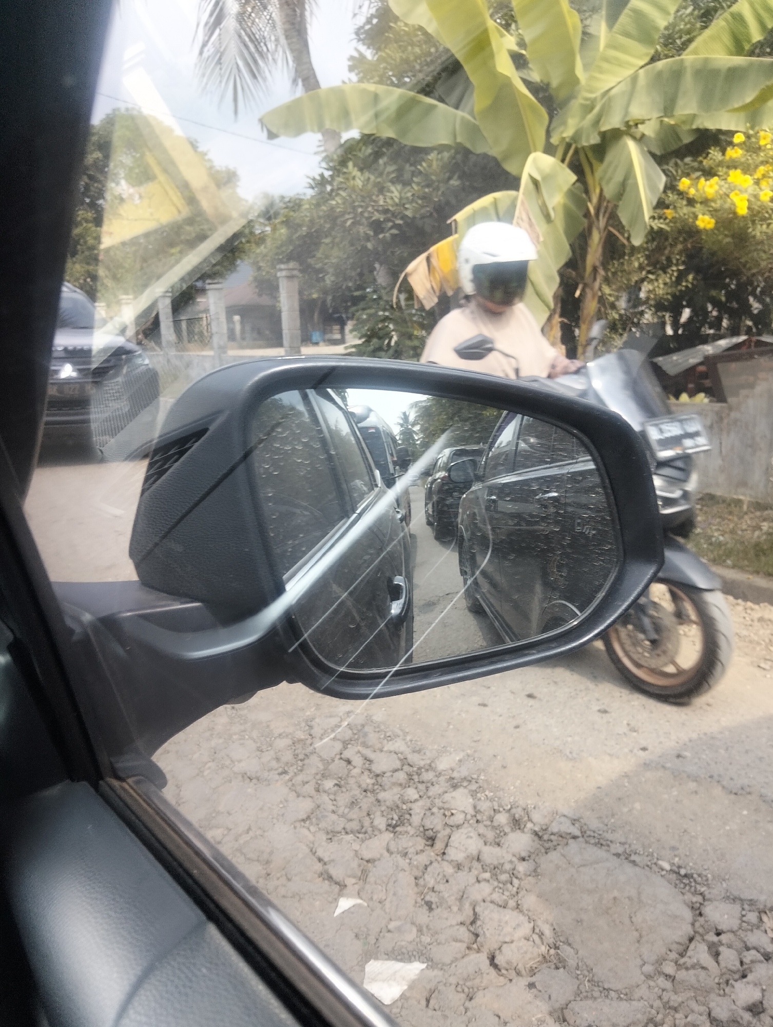 The image captures a scene from the perspective of a car driver in Teupinreudeup, Indonesia, looking into the side mirror and out the window. In the side mirror, two black cars are visible in traffic, with raindrops on the mirror's surface. Outside the car, a person wearing a white helmet and a light-colored shirt rides a motorcycle. Behind the motorcycle rider, a banana tree with large green leaves and yellow flowers is prominent. The ground is a dirt road with patches of asphalt. The sky is overcast. The overall environment suggests a rural or semi-urban setting.