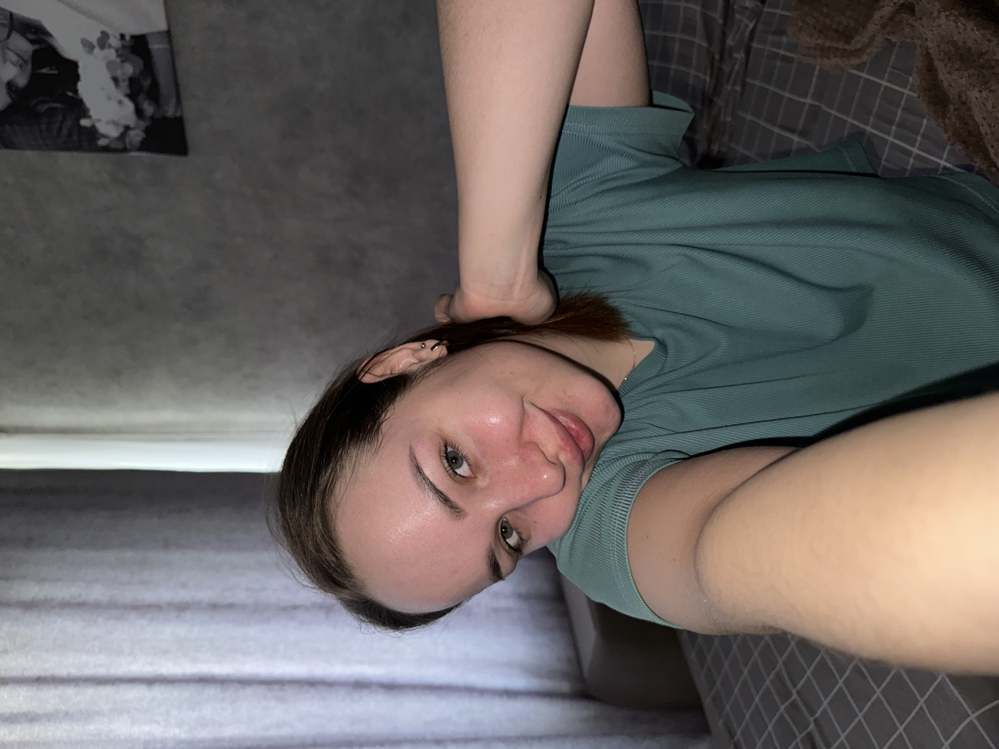 A woman is lying on a bed. She is fair-skinned with brown hair and green eyes. She is wearing a green t-shirt. One of her arms is raised over her head. Behind her are gray and brown bed linens. A gray wall is visible to her left, as well as a piece of artwork. The location is unknown, although potentially in the city of Chaikovsky, Russia.