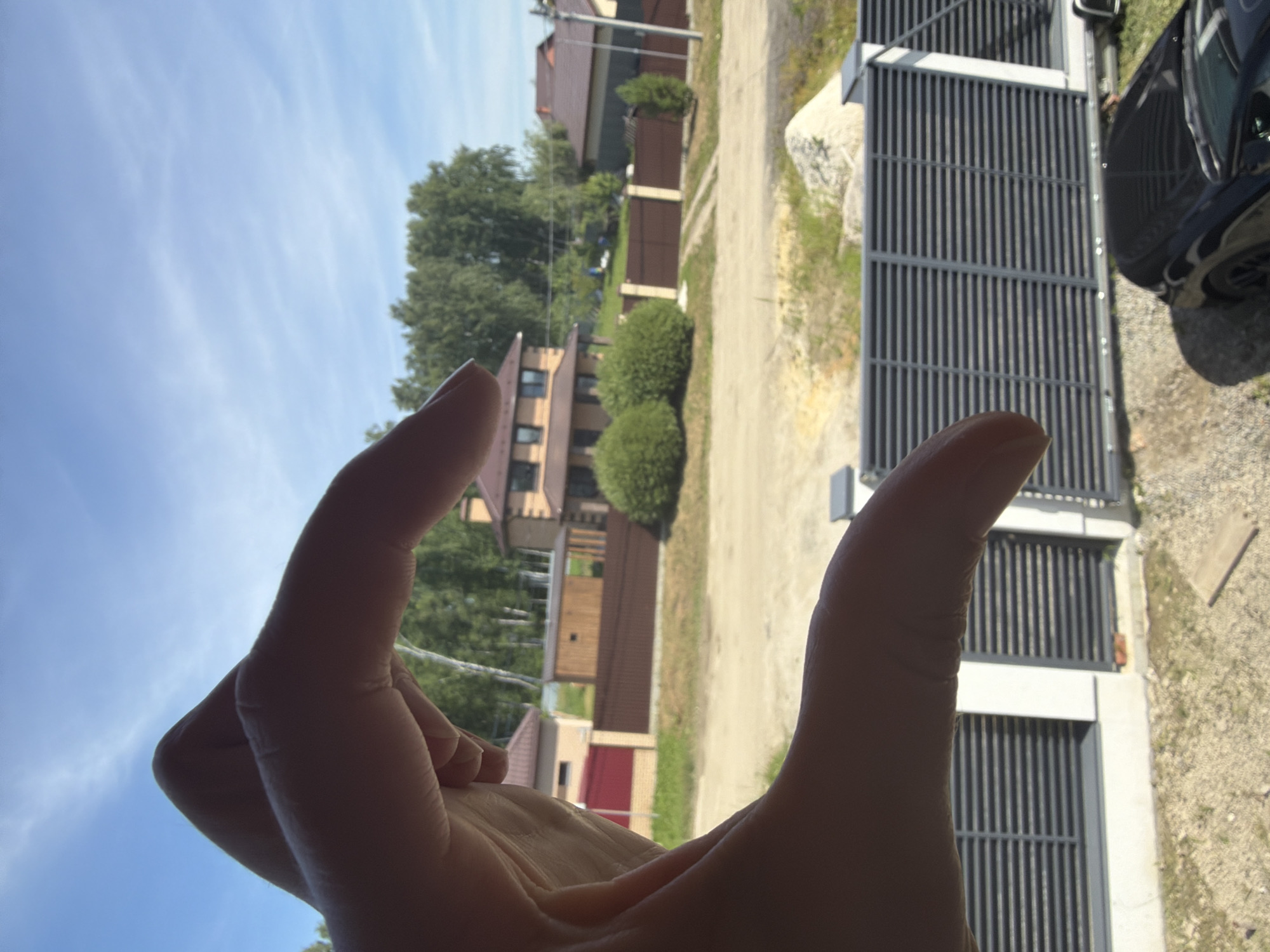 This media file features a close-up of a person's hands framing a view of a suburban scene. The hands, one with its thumb and forefinger making a circle, are in the foreground and partially obscure the background. The scene behind the hands includes several houses with red roofs, green trees, and neatly trimmed bushes. A gravel driveway leads to a modern grey metal fence and gate. To the right, the dark body and wheel of a car are visible, suggesting a residential street or driveway. The sky is a pale blue with wispy clouds, indicating it is daytime, likely with fair weather. The overall impression is of a casual, outdoor moment, perhaps someone framing a shot or simply enjoying the view. No visible text is present in the image. The context provided places the scene in Malinovka, Russia.
