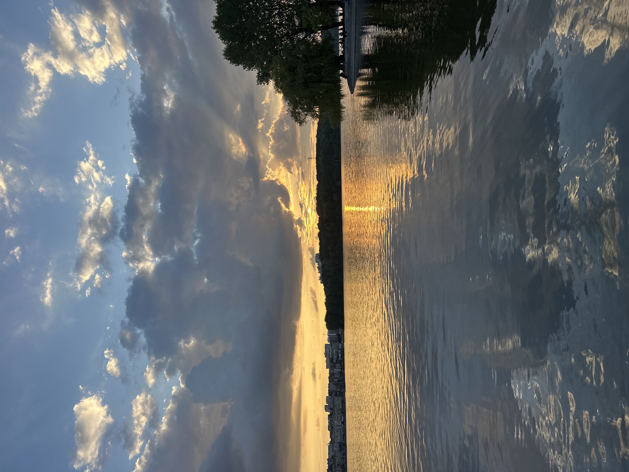 The image depicts a sunset over Ternopil Lake in Ternopil, Ukraine. The sky is a mix of blue and gray clouds, with rays of sunlight piercing through the clouds, creating a dramatic effect. The sun is low on the horizon, casting a warm golden glow across the water and the cityscape in the distance. The water is calm, with gentle ripples reflecting the sky and the surrounding landscape.
On the left side of the image, a cityscape with buildings is visible along the shore. On the right side, a lush green tree stands tall, casting a shadow on the water. Several people are seated or standing near the tree, appearing to enjoy the sunset. The overall scene is serene and picturesque, capturing the beauty of Ternopil Lake at dusk.