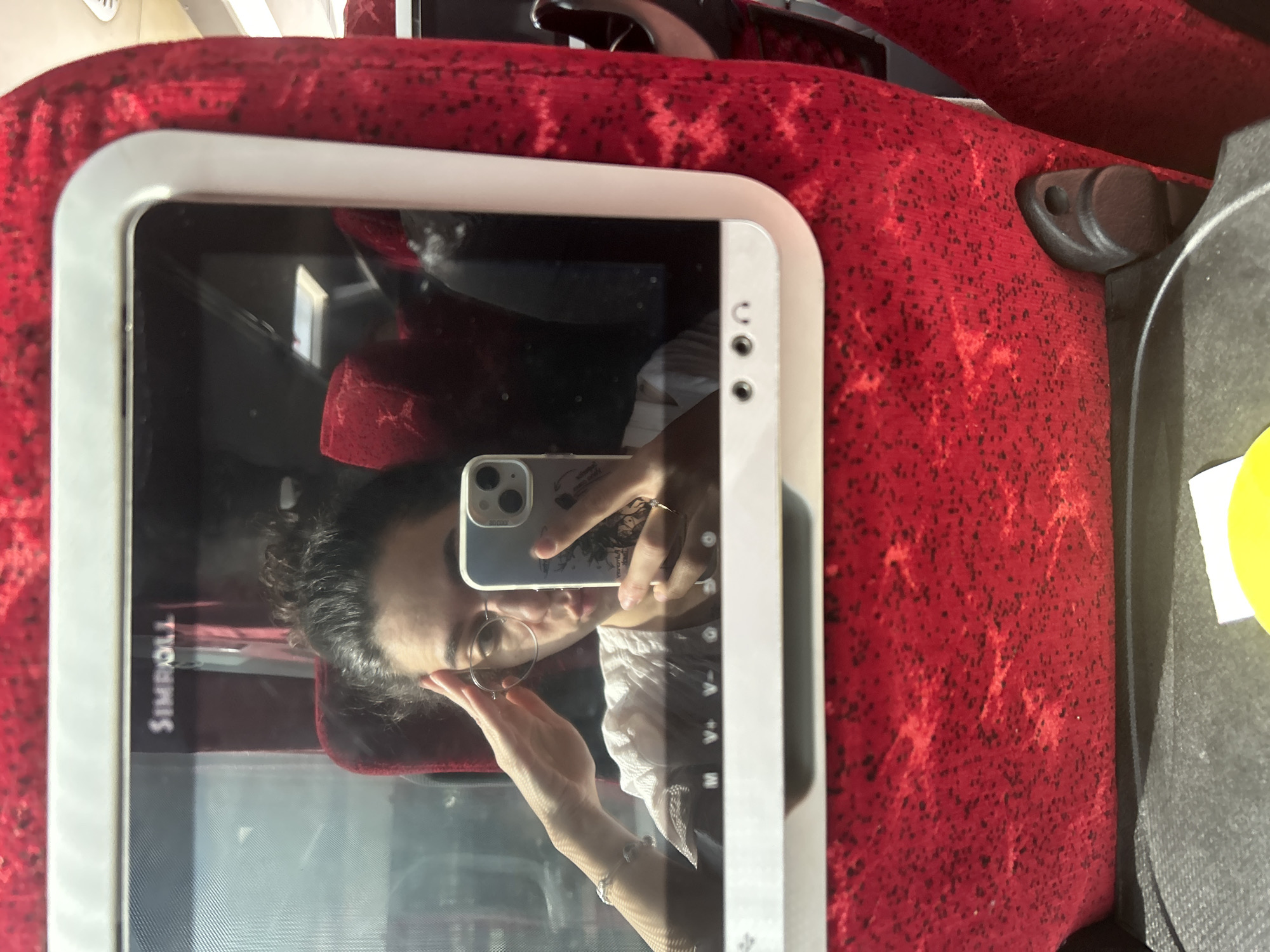 The image shows a reflection of a person taking a selfie in what appears to be a screen or a window of a public transport vehicle. The person, with dark, curly hair and wearing round-framed glasses, is holding a light-colored smartphone (likely an iPhone given the dual camera setup) to capture the image. A design or sticker is visible on the back of the phone case. The person is wearing a light-colored top with some textured detailing and a bracelet on their left wrist.
The immediate setting around the reflective screen is characterized by vibrant red fabric upholstery with a black speckled or patterned design, typical of public transport seating. The screen itself has a white/light gray bezel. To the right of the screen, a black plastic fixture, possibly part of the seat or armrest, is visible, along with a small yellow object partially peeking into the frame.
The activity is the person taking a mirror selfie. The lighting suggests it is daytime. A notable detail is the mirrored text "SIMROLL" (appearing as "110RWI S" due to reflection and image orientation) visible on the screen's surface in the reflection. The overall scene suggests the person is traveling in a bus or train in Bayrampaşa, Turkey.