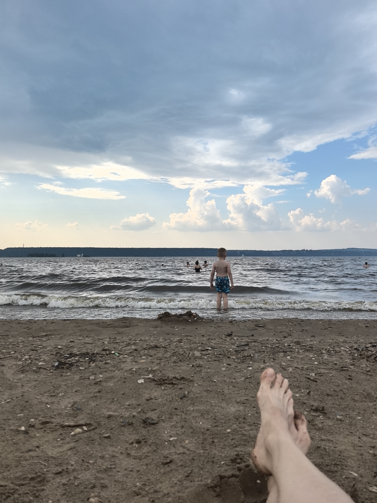 The image depicts a body of water, likely a river or lake, with a sandy beach in the foreground. The water is moderately wavy, and several people are visible swimming or wading in the distance. A young boy, seen from behind, stands at the edge of the water wearing blue patterned swim trunks. The beach is covered with sand and small debris. In the foreground, a pair of bare feet, dirty from the sand, are resting on the beach, suggesting the perspective of someone relaxing on the shore. The sky above is partly cloudy with a mix of blue sky and grey clouds. The background shows a tree line on the horizon. The scene is likely a public beach on a warm day.