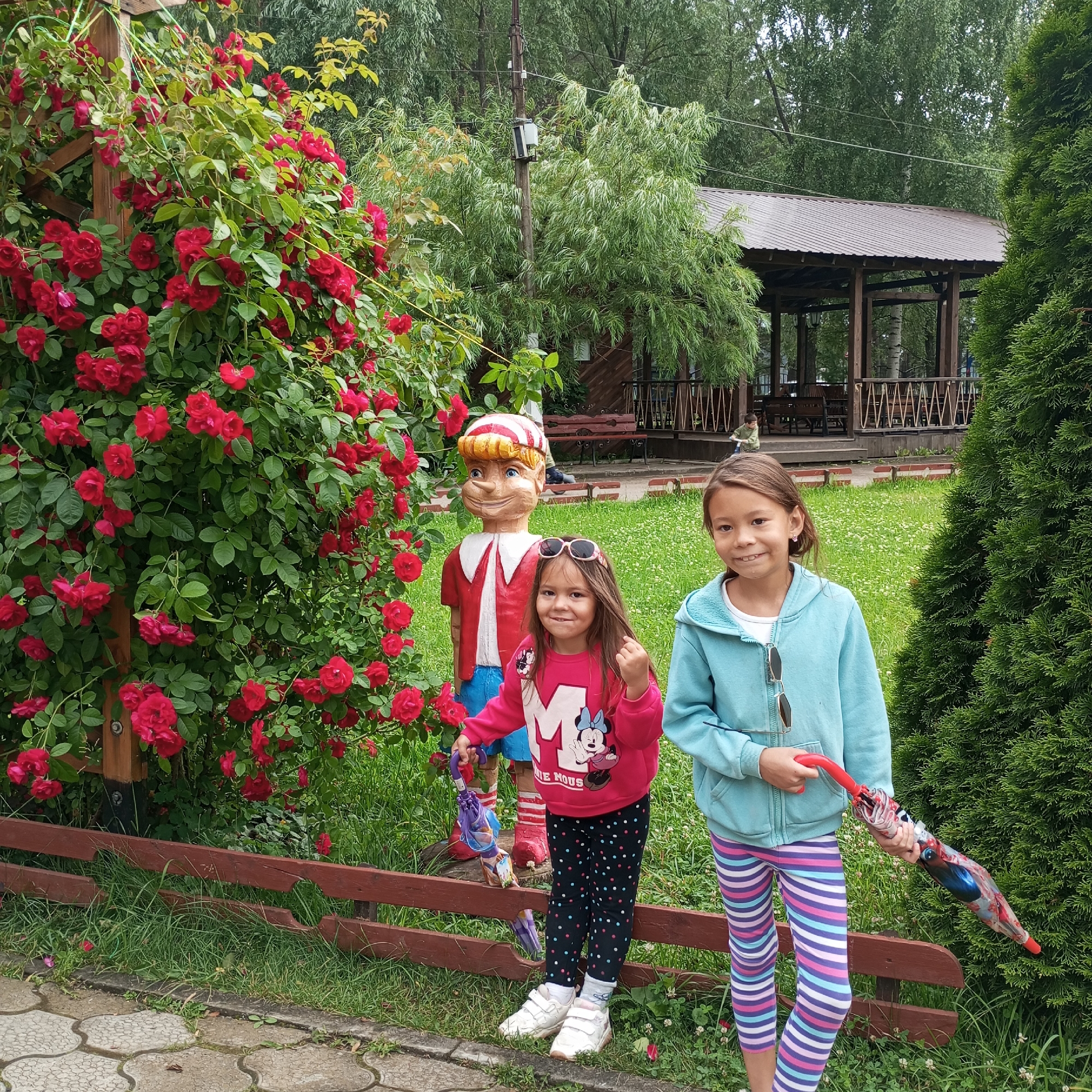 Two young girls are posing in front of a rose bush and a wooden statue of Pinocchio. The girl on the left is wearing a pink sweatshirt with a "Minnie Mouse" graphic and black pants with colorful polka dots. She is holding a small purple umbrella. The girl on the right is wearing a light blue hooded jacket and striped leggings. She is holding a colorful umbrella. The Pinocchio statue is dressed in a red jacket with a white collar and blue pants. Red roses are in full bloom on the bush. In the background, there is a wooden gazebo with a brown roof, trees, and a fence. The scene appears to be outdoors in a park or garden on a cloudy day. The girls seem to be happy and enjoying their time. The text "MINNIE MOUSE" is visible on the pink sweatshirt. The location is Chaikovsky, Russia.
