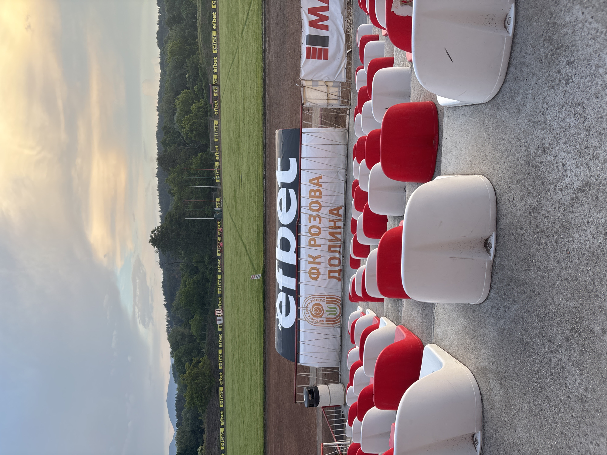 An outdoor sports stadium or football pitch, located in Kazanlak, Bulgaria, is captured in the late afternoon or early evening light. The image shows rows of empty, slightly worn red and white stadium seats in the foreground, facing a well-maintained green grass field. A dirt track borders the field.
In the midground, various advertising banners are visible. Along the perimeter of the field, multiple yellow banners display the text "efbet". Behind what appears to be an end zone or sideline, a large black and white banner prominently features "efbet" alongside "ФК РОЗОВА ДОЛИНА" (FK Rozova Dolina), which translates to FC Rose Valley, indicating the local football club. Another white banner beneath this features a circular logo and the text "КАЗАНЛЪК" (Kazanlak), confirming the specific location. A red and white banner with a large "M" or "MA" logo is also present near the seating area.
The background features lush, forested hills and distant mountains, emphasizing the stadium's natural setting. The sky above is partly cloudy, with hues ranging from soft yellows and oranges on the left, suggesting a sunset or sunrise, to cooler grey-blue tones on the right, indicating varied atmospheric conditions. No people are visible, lending a quiet, anticipatory atmosphere to the empty stadium.