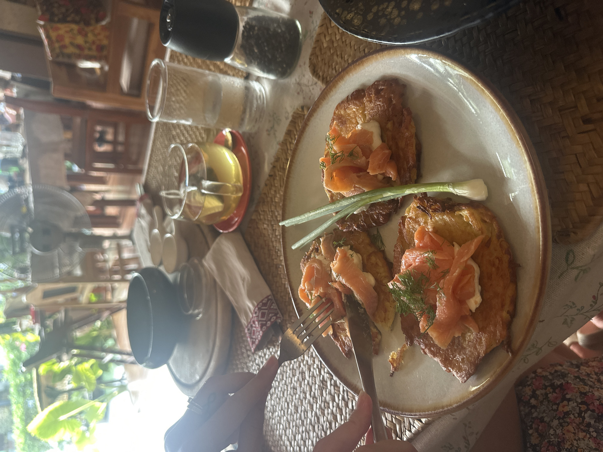 The image shows a plate of food consisting of what appears to be potato pancakes topped with salmon, a white sauce (possibly cream cheese or sour cream), dill, and spring onion. The plate rests on a woven placemat on a table covered with a tablecloth. A person's hands are visible, holding a fork and knife, poised to cut into the food. In the background, there are salt and pepper shakers, a glass of liquid, and other dining items. The setting suggests a meal is being enjoyed in a restaurant or at home. It is plausible this image was taken in Baan Chaweng Noi, Thailand, although there are no elements in the image that directly confirm the location.