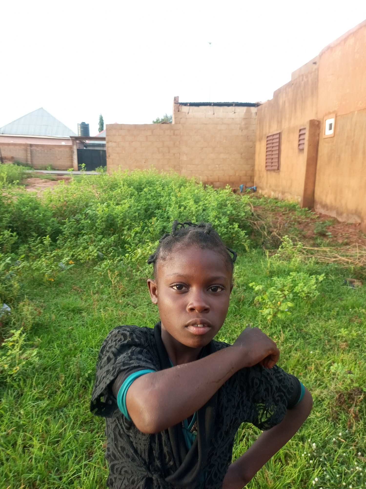 A young girl with dark skin and braided hair stands in a grassy area in front of a building. She is wearing a black lace top with a teal band visible on her arm. Her right arm is bent, and her hand rests on her bicep, while her left arm is not visible. She is looking directly at the camera with a neutral expression.
The background consists of overgrown green vegetation and a building made of light brown bricks. The building has a flat roof and a window with bars, as well as a smaller rectangular window. To the left of the building, another structure with a peaked roof is partially visible. The sky above is a pale, overcast white, suggesting it might be dawn or dusk, or a cloudy day.
The setting appears to be outdoors, possibly in a residential area, given the presence of buildings. The overall atmosphere is casual and informal. There is no visible text in the image. The location is identified as Tempure, Nigeria.