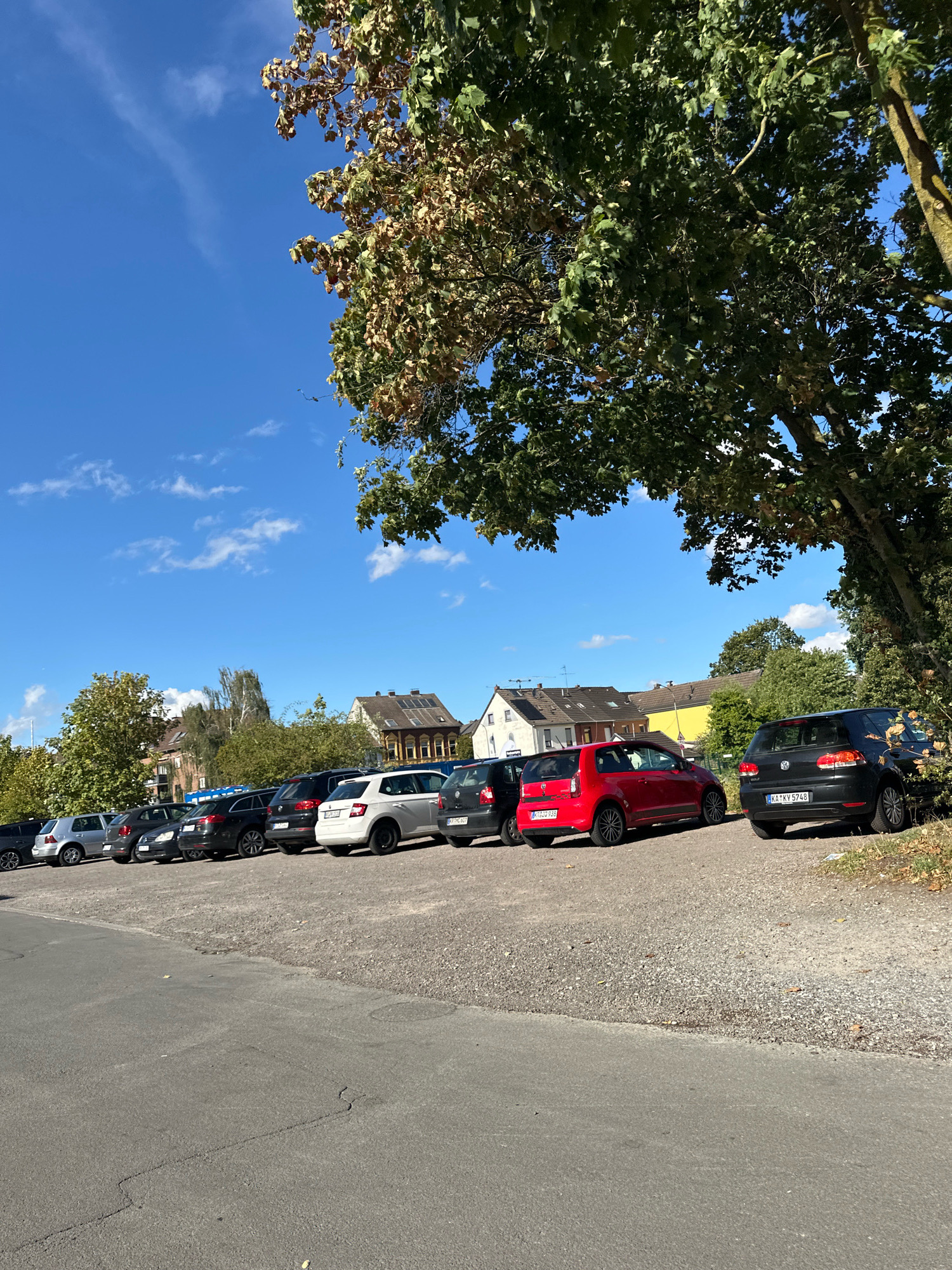The image depicts a parked row of vehicles in Krefeld, Germany, under a clear blue sky with scattered clouds. The cars are parked on a gravel surface adjacent to a paved road. The scene is bordered by trees, with a large tree in the foreground casting shadows. In the background, residential buildings with visible rooftops are present. The cars visible include a black Volkswagen Golf with license plate "KA-KY 5748," a red Volkswagen Up with license plate "KA-ZU 939," a white Skoda Fabia, and several other dark-colored cars. The environment appears to be an outdoor parking area in a suburban or residential setting within Krefeld.