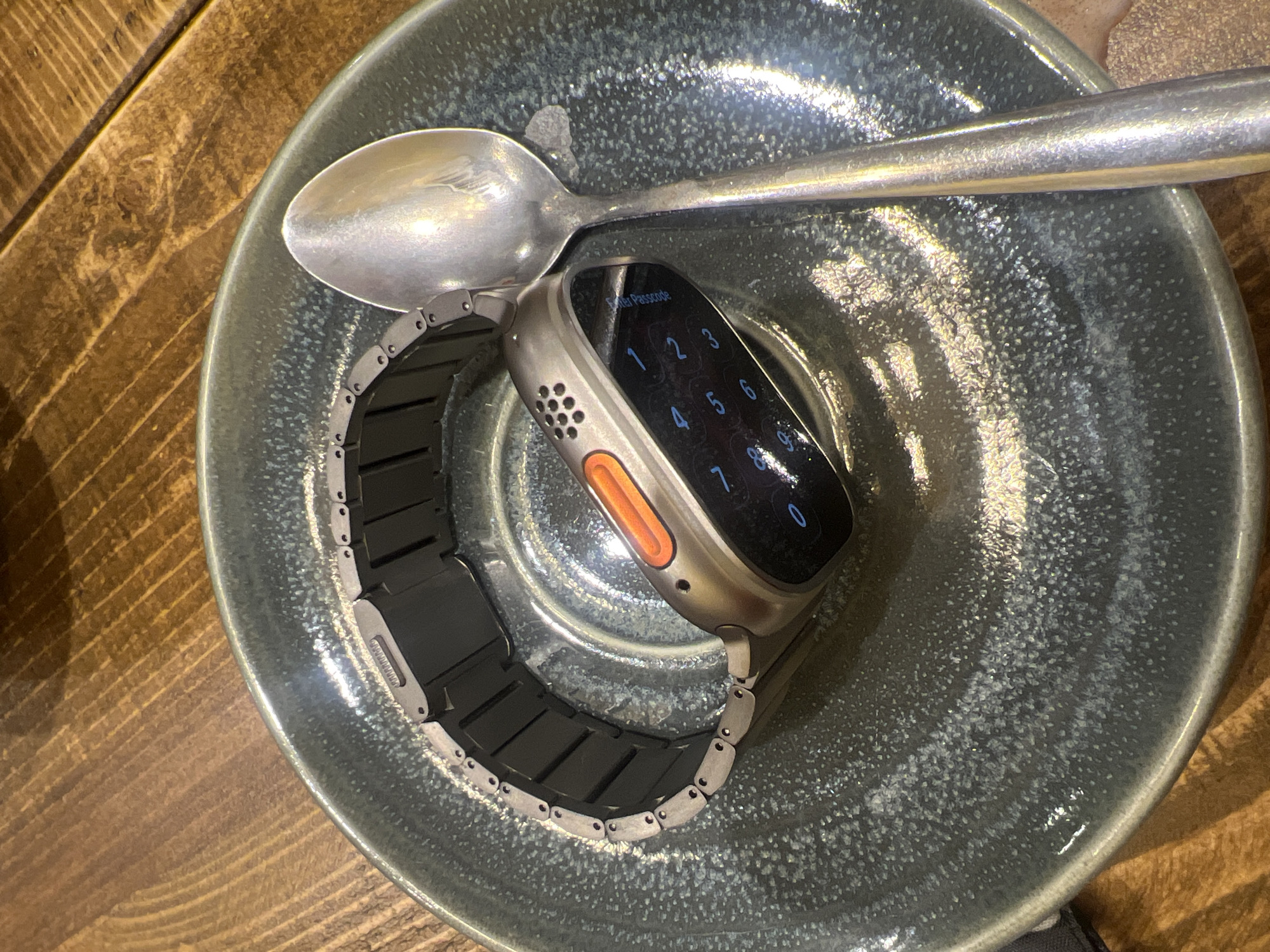 The image shows a silver Apple Watch with a black and silver band in a speckled blue ceramic bowl. A silver spoon rests across the top of the bowl. The watch display shows a number pad, indicating a passcode is required. The bowl sits on a wooden surface with visible wood grain. The location is not discernible from the image. The only city/region in the UK mentioned is Mole Valley, which is likely where the image was taken, but the environment is not specified.