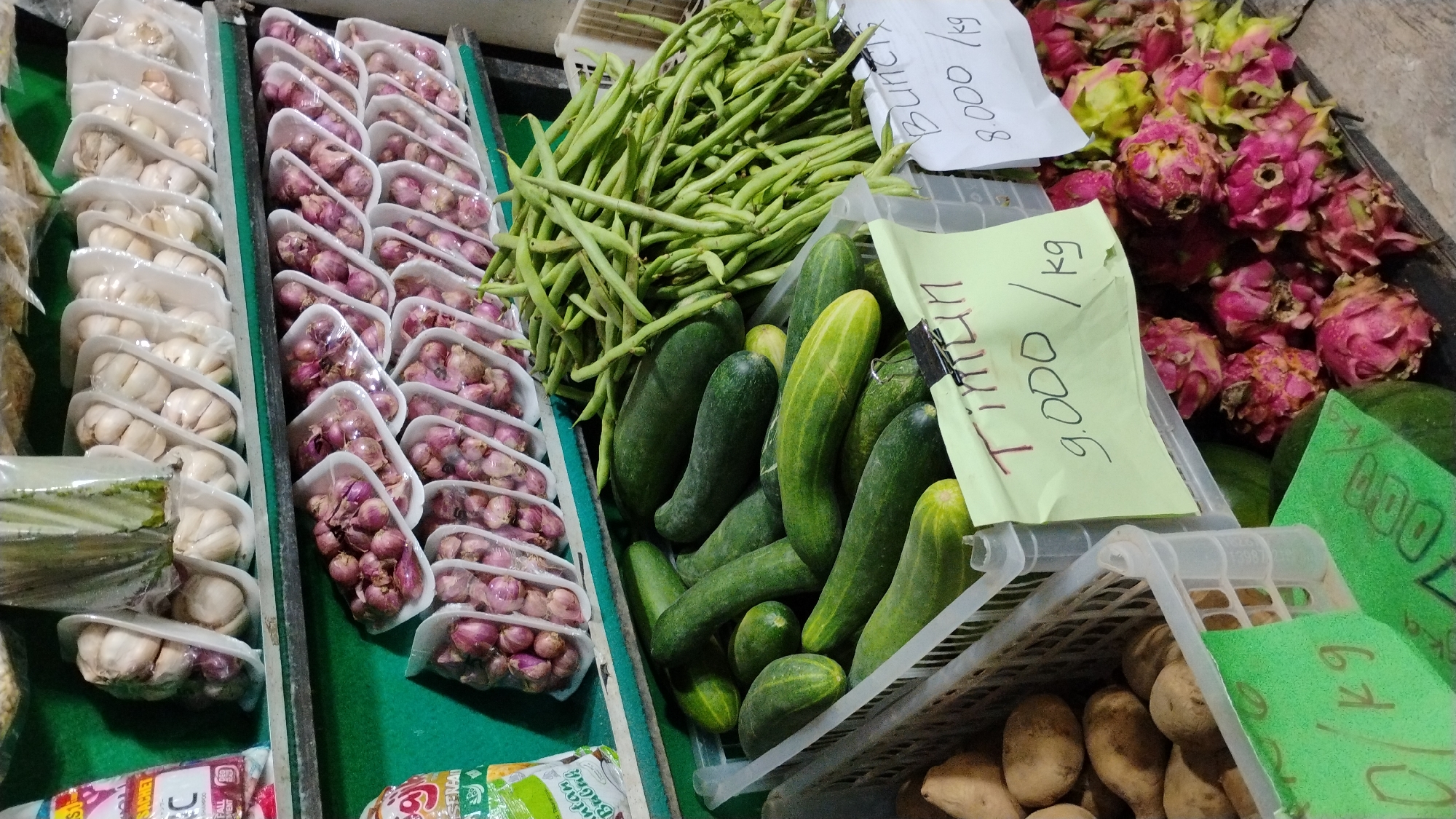 This image depicts a vibrant market stall overflowing with fresh produce. The main subjects are a variety of vegetables and fruits, including rows of garlic bulbs neatly packaged in white plastic containers, clusters of red shallots also in clear plastic packaging, a large pile of green beans, and a collection of green cucumbers. In the background, bright pink dragon fruits are prominently displayed.
The setting appears to be an outdoor market, likely in Banjarsari, Indonesia, suggested by the text on the price tags. The overall scene is busy and colorful, characteristic of a local produce market. There are no people visible in the image, so the focus is entirely on the merchandise.
Several price tags with handwritten text are visible, providing details about the products. One tag in white paper reads "BUNCIS 8.000/kg," indicating green beans priced at 8,000 per kilogram. Another tag, a light yellow sheet, displays "Tintin 9.000/kg," possibly referring to a type of cucumber or another vegetable at 9,000 per kilogram. A green tag shows "7000" and another partially visible green tag reads "kg". There are also partial labels from product packaging, one of which includes the word "Sachet" and another that appears to be from shampoo packaging.
The lighting suggests it could be daytime, with no specific indications of weather conditions. The arrangement of the produce implies a recent delivery or restocking, creating a sense of abundance and freshness. The image captures the essence of a bustling marketplace where fresh ingredients are readily available.