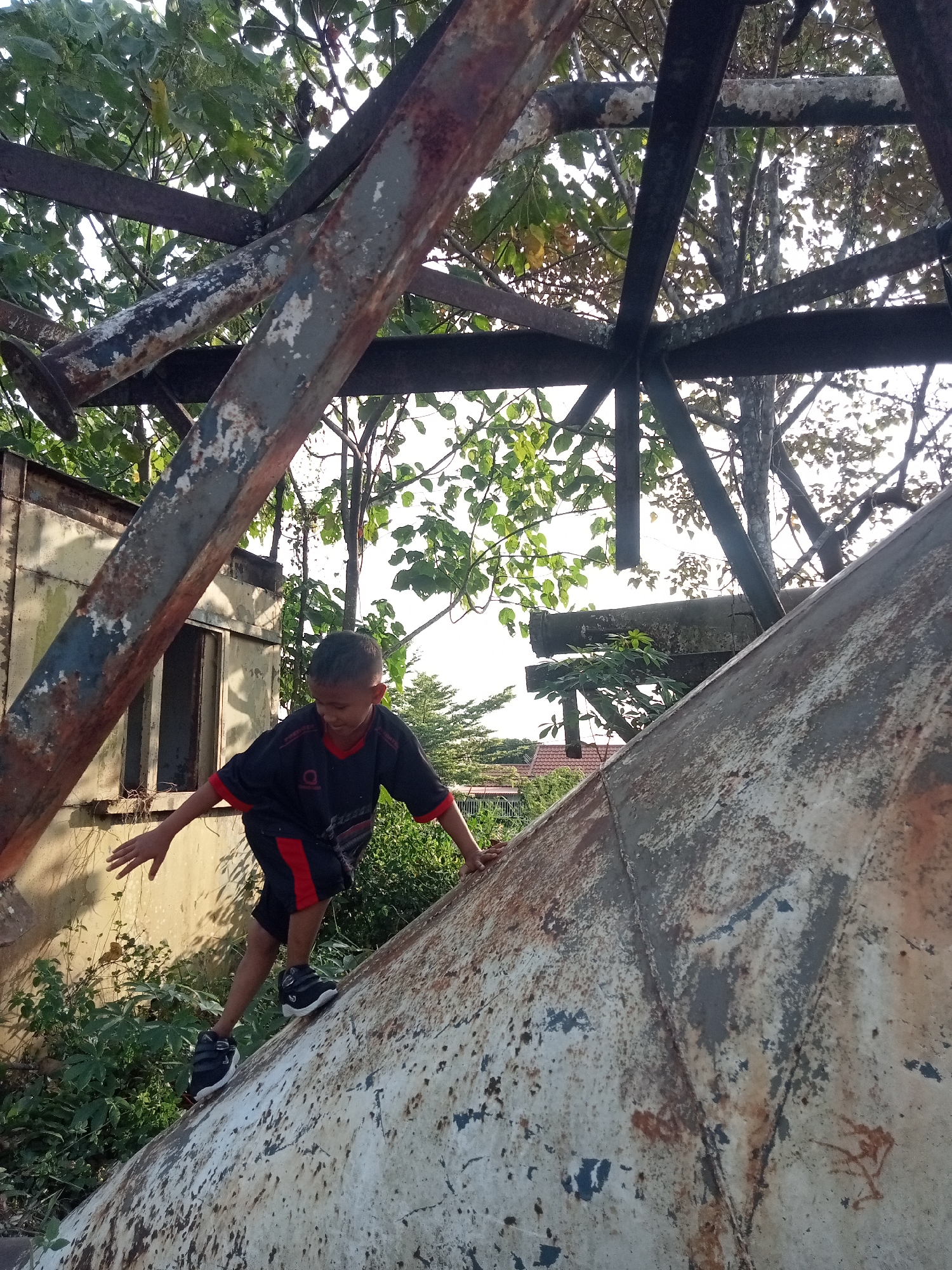A young boy with dark hair, wearing a black t-shirt with red accents and black shorts, is climbing on a large, weathered, conical metal structure. The structure appears to be made of rusted metal, with patches of peeling paint. The boy is positioned on the sloping side of the cone, with his right hand reaching out for balance and his left hand gripping the surface. He is wearing dark sneakers with white soles.
The environment surrounding the metal structure is overgrown with lush green vegetation and trees. Behind the boy and the structure, there is a dilapidated building with a yellow-painted exterior and an empty window frame. The background shows more trees and a glimpse of a residential area with red-tiled roofs, suggesting an urban or semi-urban setting. The lighting indicates it is daytime, with sunlight filtering through the trees, creating shadows.
The scene is located in Teluk Kenidai, Indonesia.