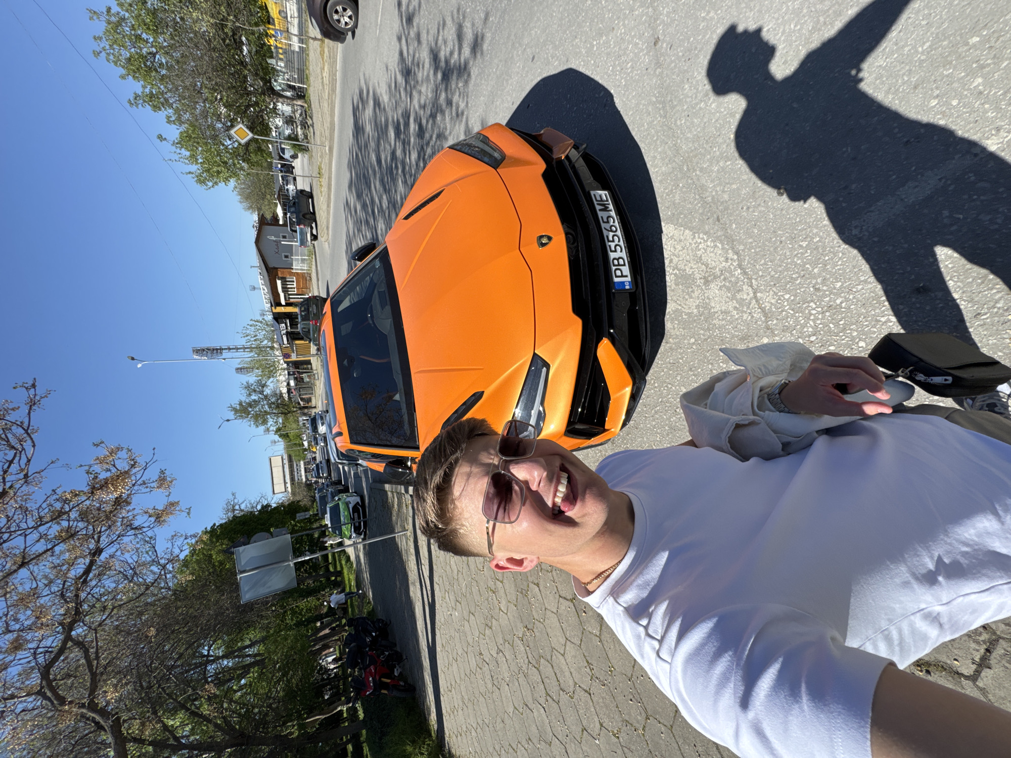 The image is a selfie of a young man standing next to an orange Lamborghini in Plovdiv, Bulgaria. The man is smiling and wearing sunglasses and a white t-shirt. His shadow is cast on the pavement. The car has a Bulgarian license plate. The scene appears to be on a paved street with trees, parked motorcycles, and other vehicles in the background. The sky is blue. The photo is taken during the daytime.