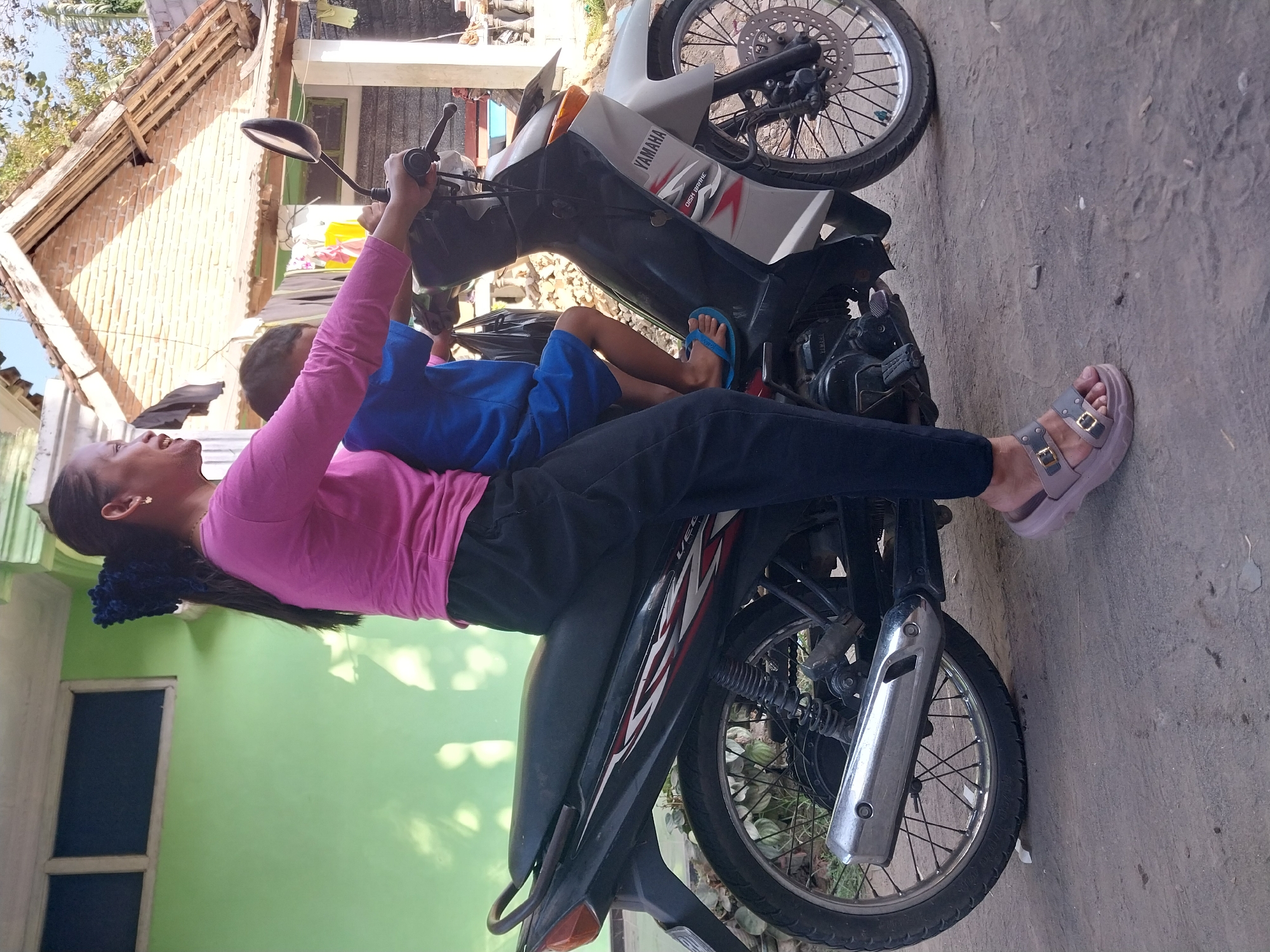 In a residential area of Nglegok, Indonesia, a woman and a child are on a Yamaha motorcycle. The woman, wearing a pink long-sleeved shirt and black pants, is seated at the front, operating the motorcycle. The child, in a blue shirt and shorts, sits in front of her. The motorcycle is parked on a dirt or gravel surface, with a green building and a brick house in the background. The scene appears to be outdoors, possibly in a yard or alleyway, given the presence of vegetation and the general state of the buildings. The lighting suggests it is daytime.