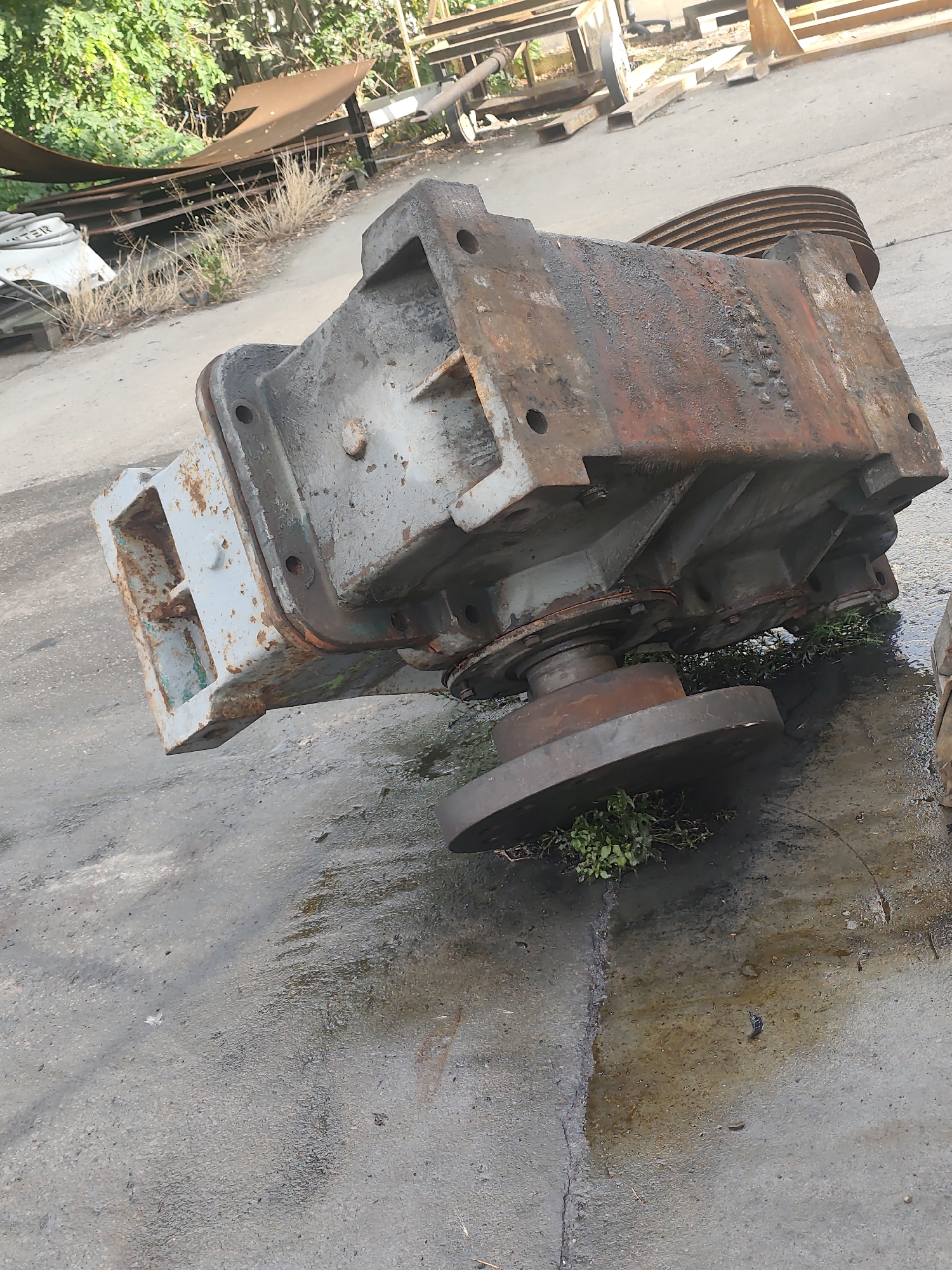 In the city of Police, Poland, a large, heavily rusted piece of industrial machinery rests on a wet, grey concrete surface. The machinery, a complex assembly of metal parts, is positioned at an angle, with its main body appearing to be a large, rectangular block. A pulley system with multiple grooves is visible at the top of the block. A circular, flat metal disc is attached to the underside of the machinery, resting on a circular shaft. Some green vegetation is growing through cracks in the concrete beneath the disc. The surrounding environment appears to be an outdoor industrial or storage area, with other metal structures and vegetation visible in the background. The concrete is marked with water stains and what appears to be oil or grease.
