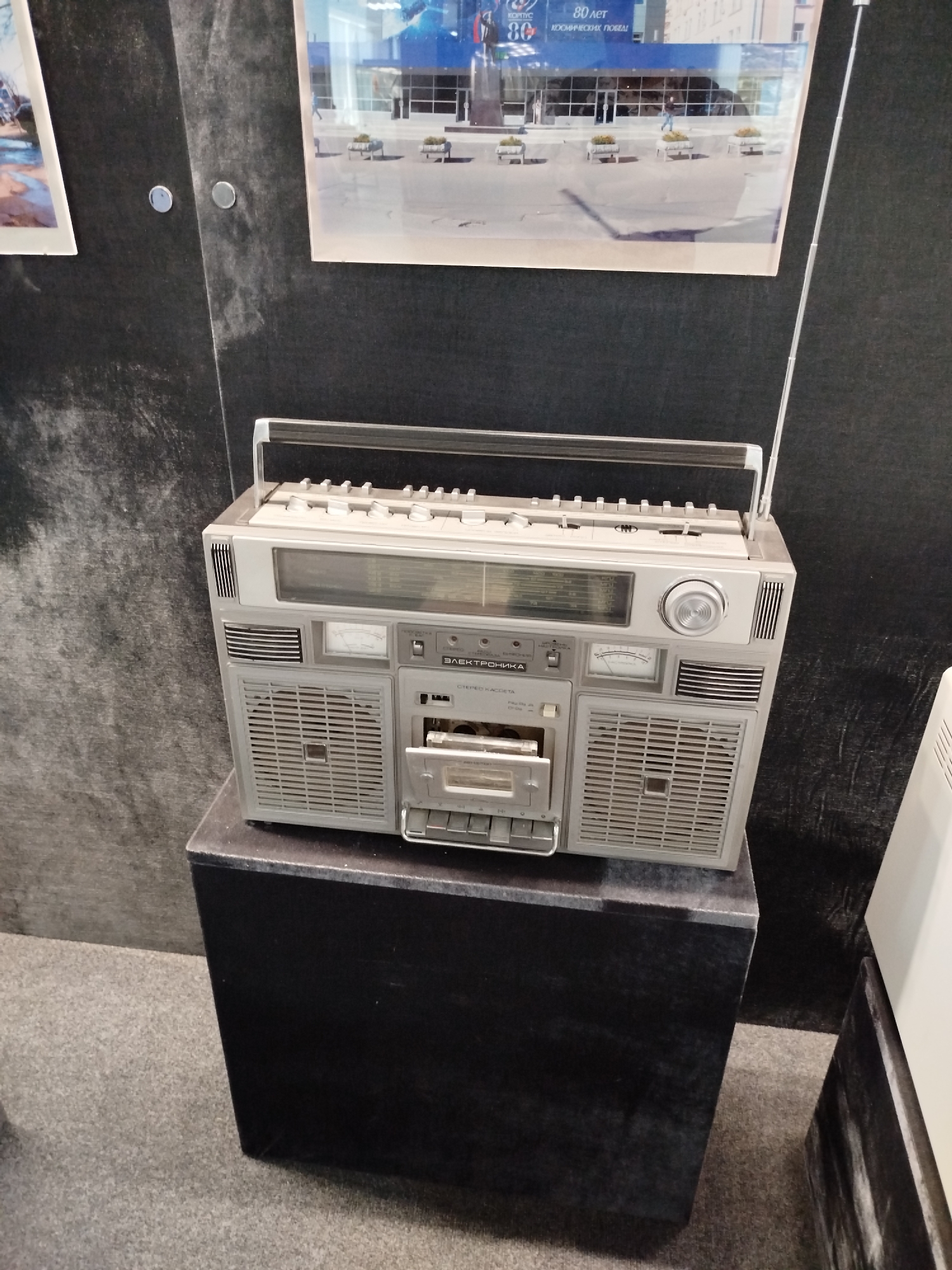This image shows a vintage boombox placed on a black pedestal. The boombox is silver and gray, with a cassette player, radio tuner, and speakers. It has a retractable antenna and a handle on top. The cassette player door is open, revealing a cassette tape. Above the boombox, a framed photograph is mounted on a dark wall. The photograph appears to be of a building with a blue facade, and text is visible on it, including "80 лет" and "КОСМИЧЕСКИХ ПОБЕД". The scene is indoors, likely in a museum or exhibition, as suggested by the display and the presentation of the items. The lighting is neutral, and there are no strong indicators of time of day or weather. The overall mood is one of nostalgia and appreciation for retro technology.