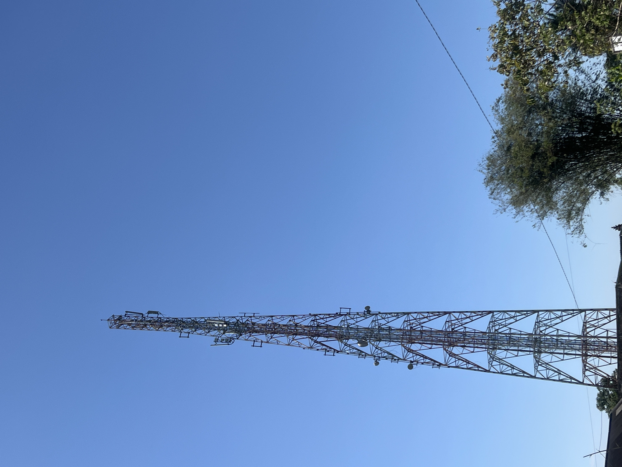 A tall, lattice telecommunications tower stands against a clear, bright blue sky in Temanggung, Indonesia. The tower, constructed of metal girders, has multiple antenna arrays attached to its structure at various heights. Below the tower, a lush green canopy of trees, including bamboo, is visible. The lower portion of the image shows the dark roofs of some buildings, suggesting an urban or semi-urban setting. A thin wire stretches diagonally across the upper right portion of the frame. The scene is illuminated by natural daylight.