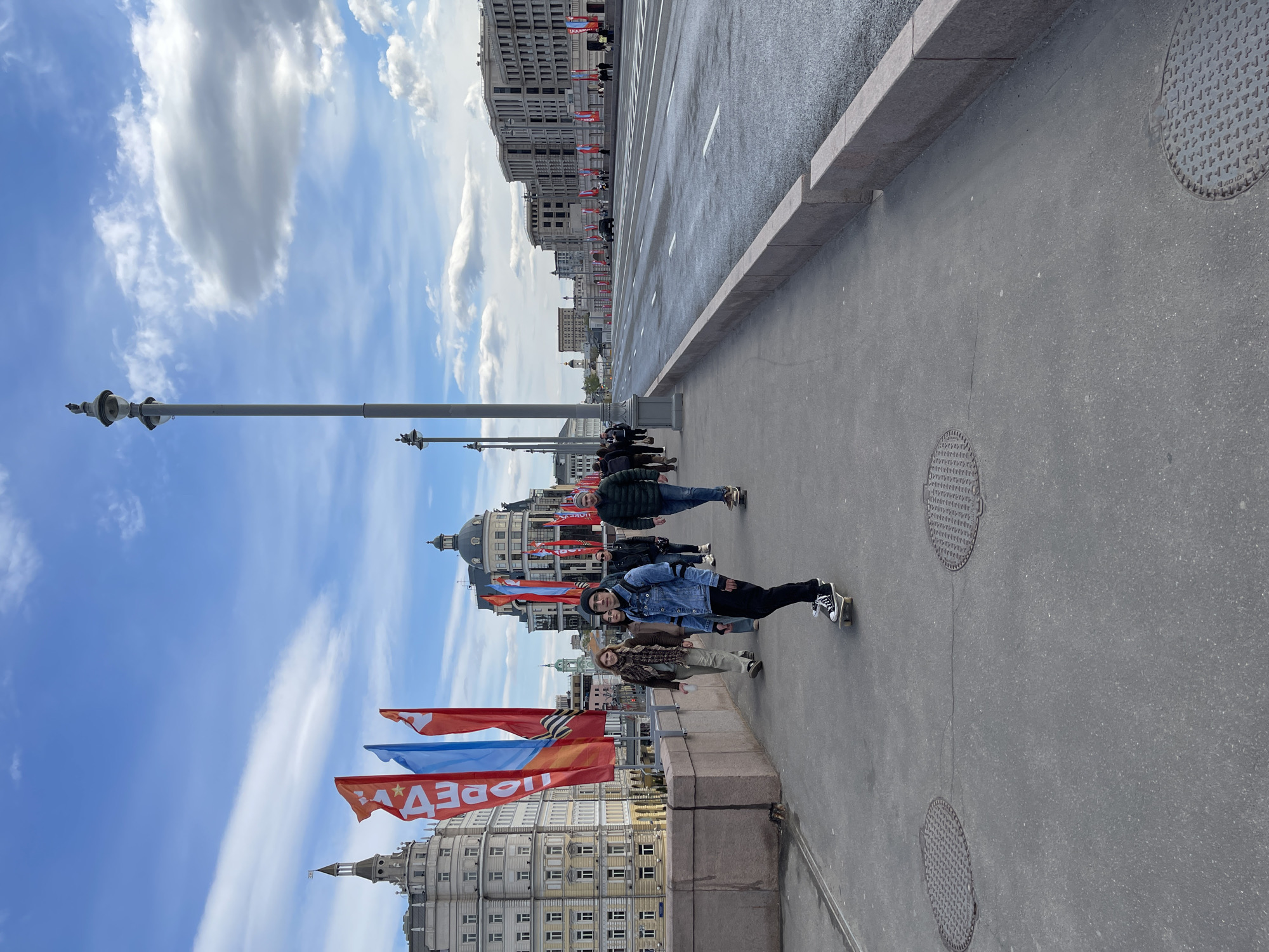Here's a description of the image:
The image shows a bridge in Moscow, Russia with people standing on the sidewalk. There are three people in the foreground. The person in the center wears a blue denim jacket, black pants, and black-and-white sneakers. They are turned towards the camera. To their left, a person in a brown jacket and light-colored pants stands next to the ledge. To their right is a person in a black jacket and blue pants.
In the background, there is a road leading towards the city center. Buildings line the street, adorned with flags. The flags have a white inscription. The sky above is blue with scattered white clouds. Light poles stand along the bridge. The road surface appears wet. Round metal manhole covers are visible in the sidewalk.