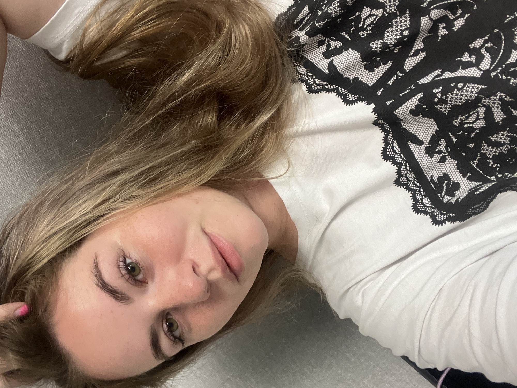 A young woman with blonde hair and light skin is lying down, looking at the camera. She is wearing a white shirt with black lace detailing on the upper right side. The background appears to be a gray surface. There is no indication in the image that indicates the location is the city of Chaikovsky, Russia.