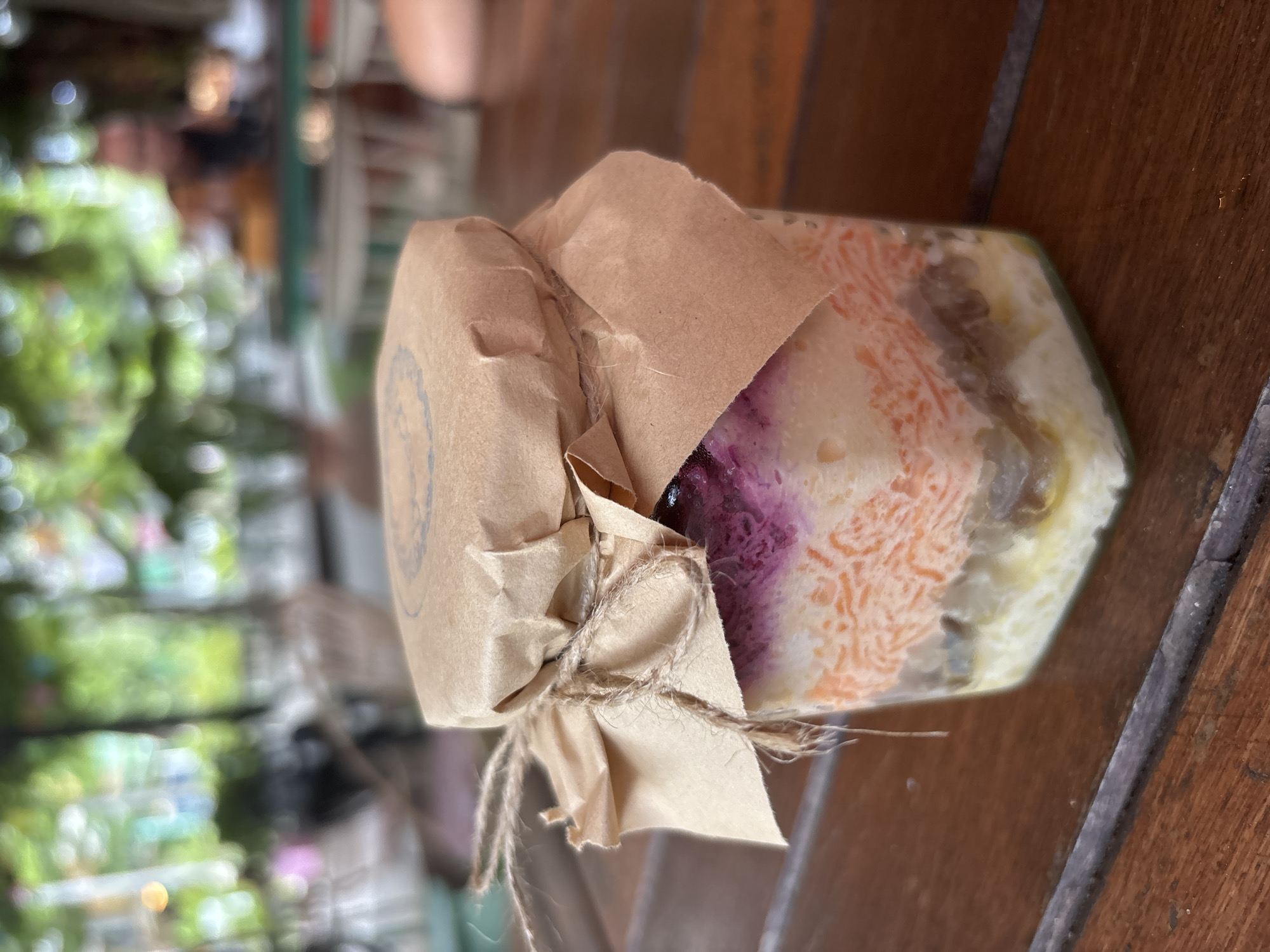Captured outdoors in บ้านบางรักษ์, Thailand, the image focuses on a glass jar filled with layered food. The contents range in color from purple to orange to white, suggesting a variety of ingredients. A brown paper wrap, tied with twine, partly obscures the jar. The jar rests on a weathered, brown wooden surface. An out-of-focus cityscape, featuring greenery and hints of buildings, provides a blurred backdrop.
