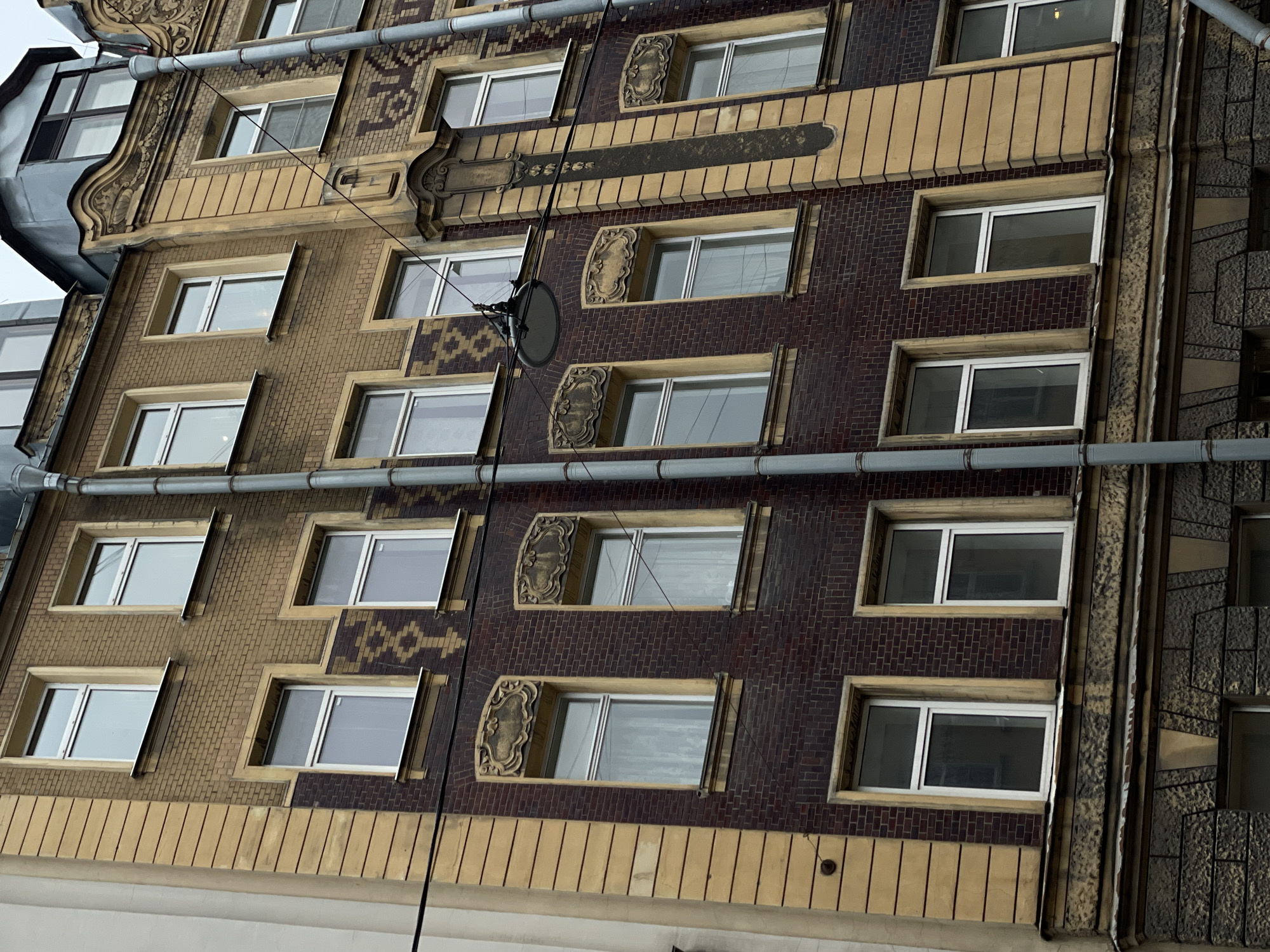 The image shows a close-up of the façade of a building in Saint Petersburg, Russia. The building has multiple stories with windows framed by decorative elements. The facade is made of brick in two different colors, tan and reddish-brown, arranged in alternating sections. A grey pipe runs vertically down the building and horizontally across each story. A satellite dish is attached to the facade between the first and second story, affixed by a cable that runs down the building. The architectural style appears to be art nouveau.