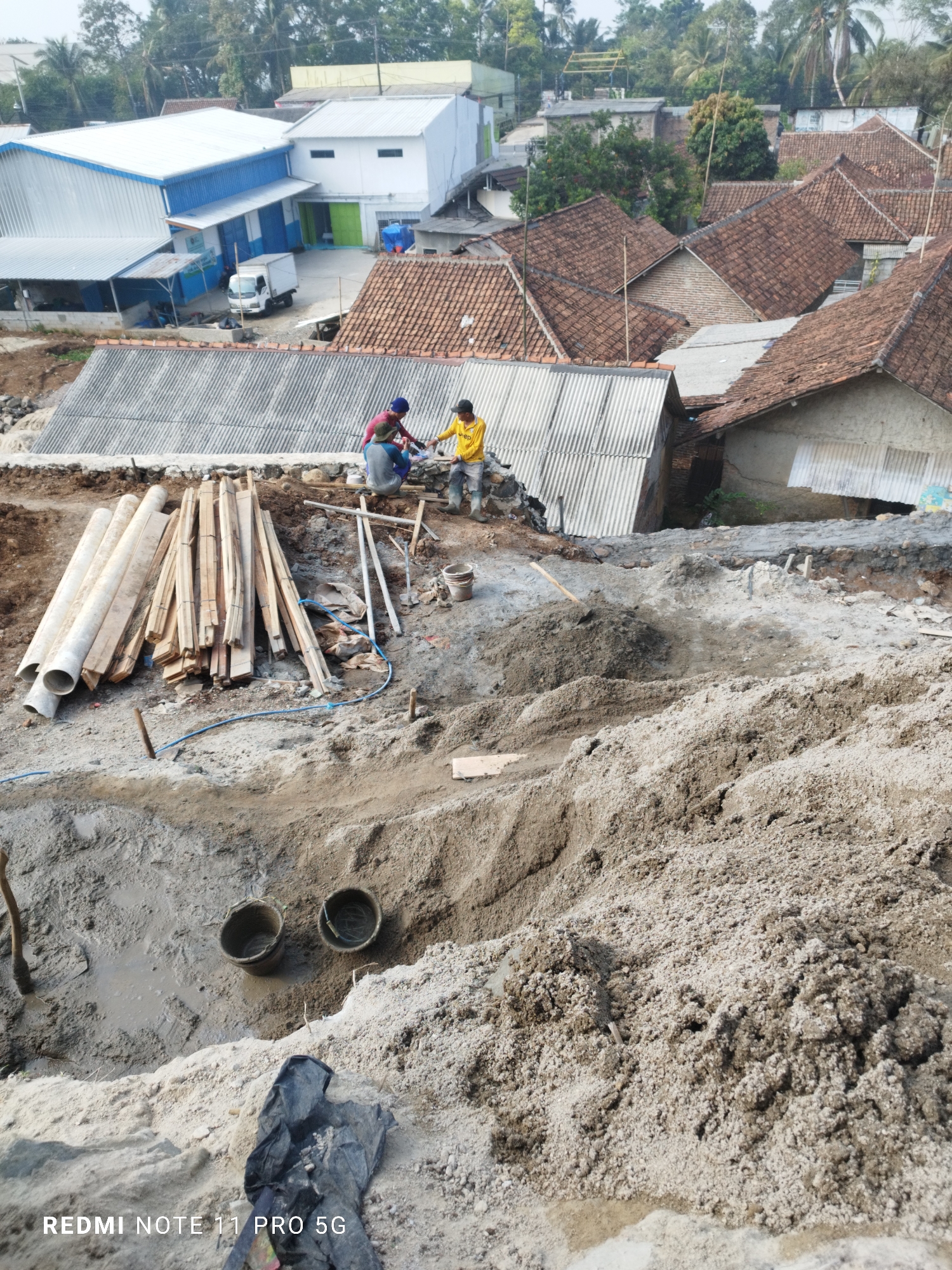 In the village of Kaduhejo, Indonesia, an aerial view captures a construction site with two workers amidst piles of sand and debris. One worker, wearing a red shirt and blue hat, is kneeling and interacting with a pile of lumber. Another worker, in a yellow shirt, stands nearby, observing the scene. Surrounding the workers are various construction materials, including pipes, wooden planks, and buckets filled with what appears to be wet cement or mud. The background reveals a residential area with houses featuring terracotta tile roofs, as well as a large industrial building with a blue and white facade. The terrain is uneven, indicating ongoing excavation or building activity.