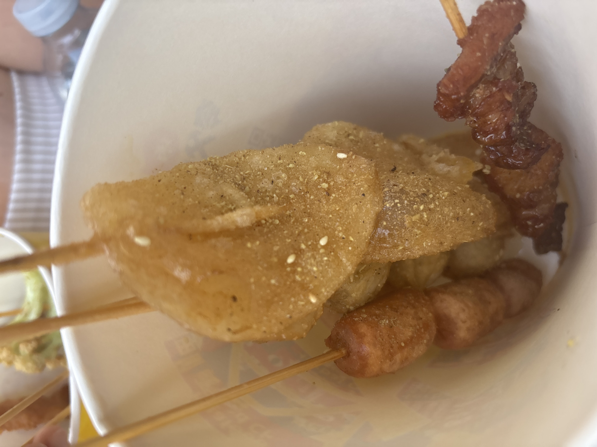 The image displays a close-up, top-down view of various skewered food items contained within a white, disposable paper cup. Dominating the center is a large, irregularly shaped, golden-brown item, likely deep-fried, coated with a visible dusting of dark spices and small white sesame seeds, impaled on a wooden skewer. To the left, a wooden skewer holds two smaller, cylindrical, reddish-brown items resembling mini sausages or hot dogs, also dusted with spices. To the right, another wooden skewer presents several small, glossy, reddish-brown pieces of grilled or caramelized meat.
The interior of the cup shows a lighter-colored liquid or sauce at the bottom. In the blurred background, partially visible elements suggest a casual dining environment: the rim of another white container holding indistinct green food (possibly vegetables), a person's upper torso clad in a white and striped garment, and the top portion of a plastic bottle with a blue cap. The lighting is bright and even. The scene depicts a selection of convenience or street food, presented for immediate consumption. This casual food service is located in the city of Milan, Italy.