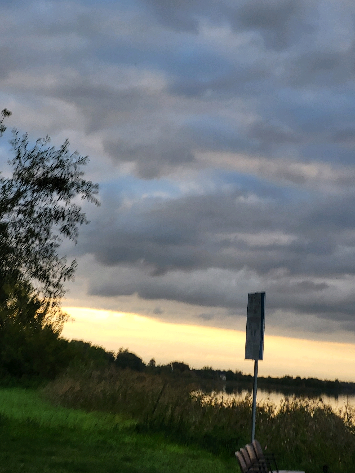 This image depicts a twilight scene near a body of water in Kalnciems, Latvia. The sky is filled with dramatic, heavy clouds, with a band of bright yellow sunlight breaking through near the horizon, illuminating the clouds from below. In the foreground, there is a grassy area and tall reeds bordering the water. To the right of the frame, a tall, rectangular signpost with a dark blue and white sign stands erect. A bench is partially visible in the lower right corner. The water reflects the light from the sky. A treeline with foliage is visible on the left side of the image. The overall atmosphere is calm and serene, characteristic of an evening by the water.