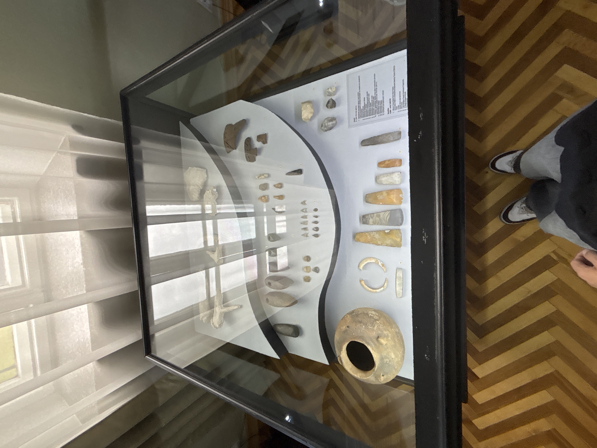 The image shows an exhibit of ancient artifacts displayed in a glass case. The artifacts include pottery, stone tools, and jewelry. The exhibit is located in a museum in Hrodna, Belarus, as indicated by a sign in the display case that reads "Hrodna". A person's legs and feet are visible in the foreground, wearing sneakers and casual clothing, suggesting they are a visitor to the museum. The exhibit is presented on multiple levels within the case, with labels providing information about the objects. The background features a window with white curtains and a wall, indicating an indoor setting. The floor is a wooden parquet with a herringbone pattern.
