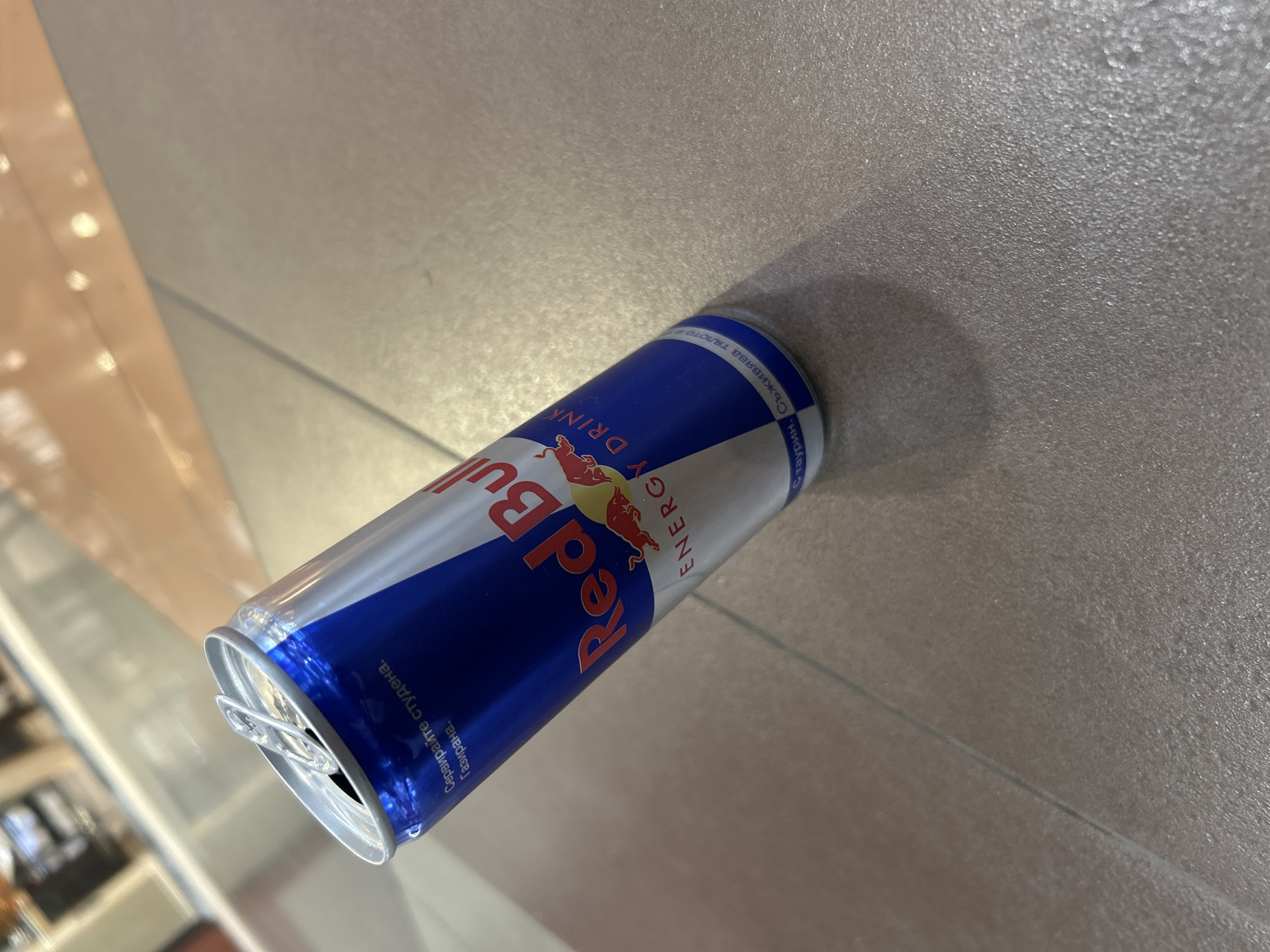 The image shows a partially opened can of Red Bull energy drink lying on a gray surface, possibly a floor or countertop. The can is blue and silver with the Red Bull logo visible. The surface appears clean and relatively smooth. The location of the image is Unknown, Unknown.