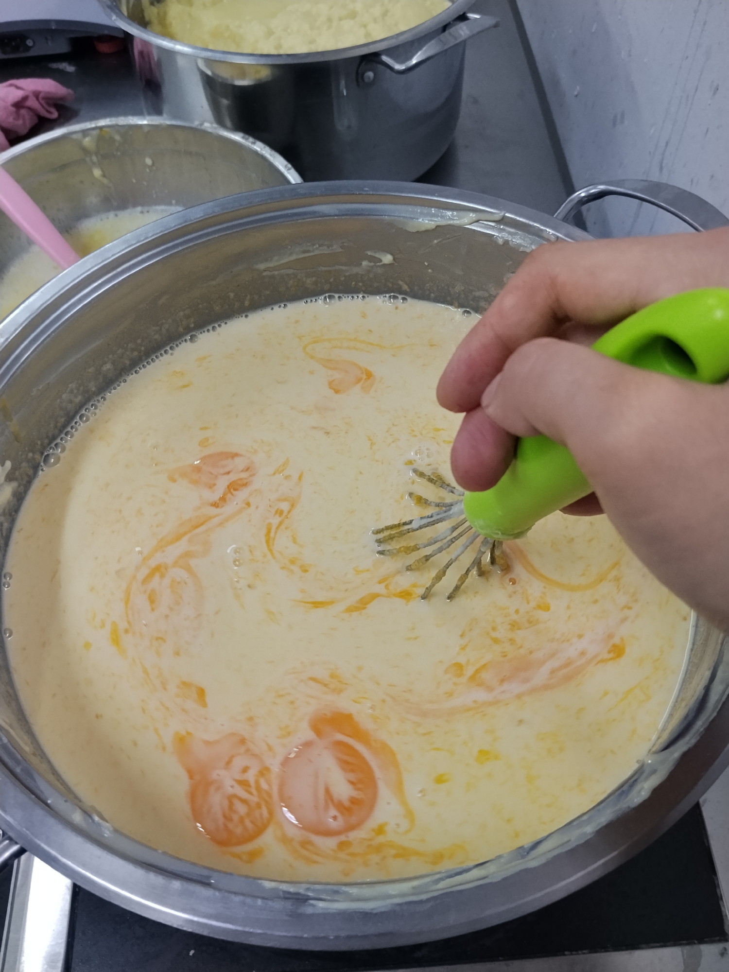 In a kitchen setting, a person's hand holds a green-handled whisk and stirs a pale yellow liquid with orange swirls in a large, stainless steel pot. The liquid appears to be a custard or sauce base, with some egg yolks visible. Another stainless steel pot containing a similar yellow substance is partially visible in the background, as is a pink spatula in a smaller metal bowl. The scene suggests food preparation.