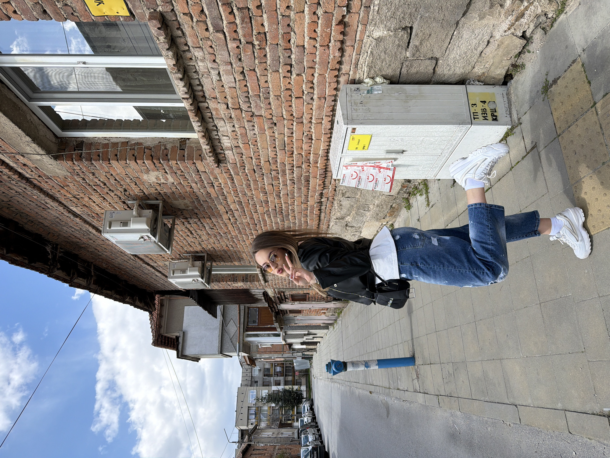 A person is standing on a sidewalk next to a building in Panagyurishte, Bulgaria. The person is wearing jeans, a white shirt, a black jacket, and white sneakers. They are also wearing sunglasses and holding up two fingers in a peace sign. The building is made of brick and has several windows and air conditioning units. There is a white electrical box with stickers on it next to the person. In the background, there is a street with cars parked on it and other buildings. The sky is blue with some clouds.