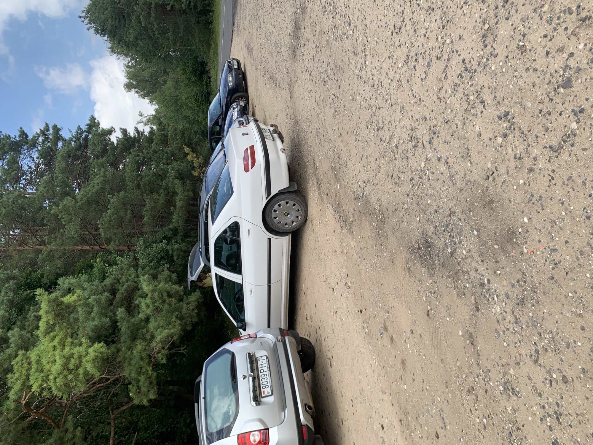 A silver Dacia Duster and a white Peugeot 406 sedan are parked side-by-side on a dirt and gravel lot at the edge of a forest. The Dacia is in the foreground, facing away from the viewer, with its tailgate open. The Peugeot is to the right of the Dacia, also facing away from the viewer. A black Volvo S80 sedan is parked further down the row of cars. The lot is bordered by a dense forest on the left and a paved road on the right. The sky is blue with scattered white clouds. The license plate on the Dacia reads "8039 PH-7 BY". The license plate on the Peugeot is partially visible and appears to read "7161 EH-5". A person in a yellow shirt is visible standing by the open tailgate of the Dacia. The scene appears to be in a rural or wooded area in the Грозаўскі сельскі Савет, Belarus.