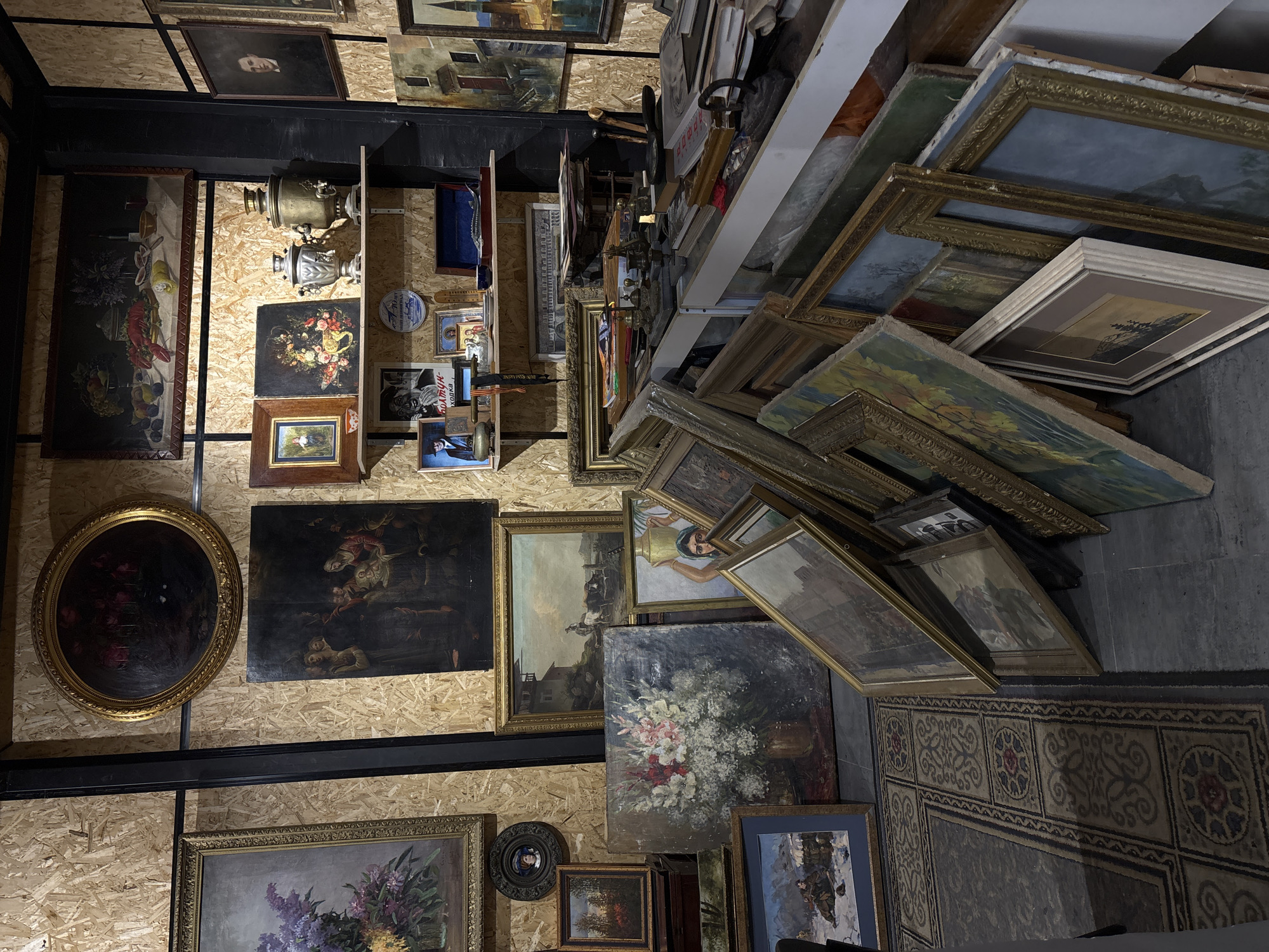 The image depicts an interior space cluttered with a large collection of framed paintings and other objects. The walls are covered with wooden panels, on which numerous paintings are hung in a somewhat haphazard arrangement. Some of the paintings depict landscapes, portraits, and still life subjects, while others are less discernible due to the low lighting. Shelves above are stacked with more framed artworks, leaning precariously. On the floor, a pile of paintings leans against the wall, some resting on a carpet with a patterned design. In the context of Saint Petersburg, Russia, this scene may represent a storage area, an antique shop, or the home of an avid art collector. The overall atmosphere suggests a dense accumulation of art and artifacts within a confined space.