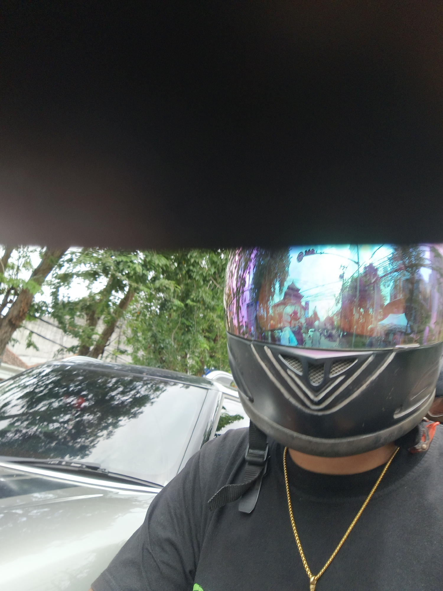 A selfie taken by a person wearing a black helmet with a reflective visor, a black t-shirt, and a gold chain. The visor reflects a street scene with traditional buildings, trees, and people. In the foreground, a silver car is visible. The scene appears to be an outdoor environment in the city of Denpasar, Indonesia, possibly a market or a festival given the crowded reflection. The overall lighting suggests it is daytime.
