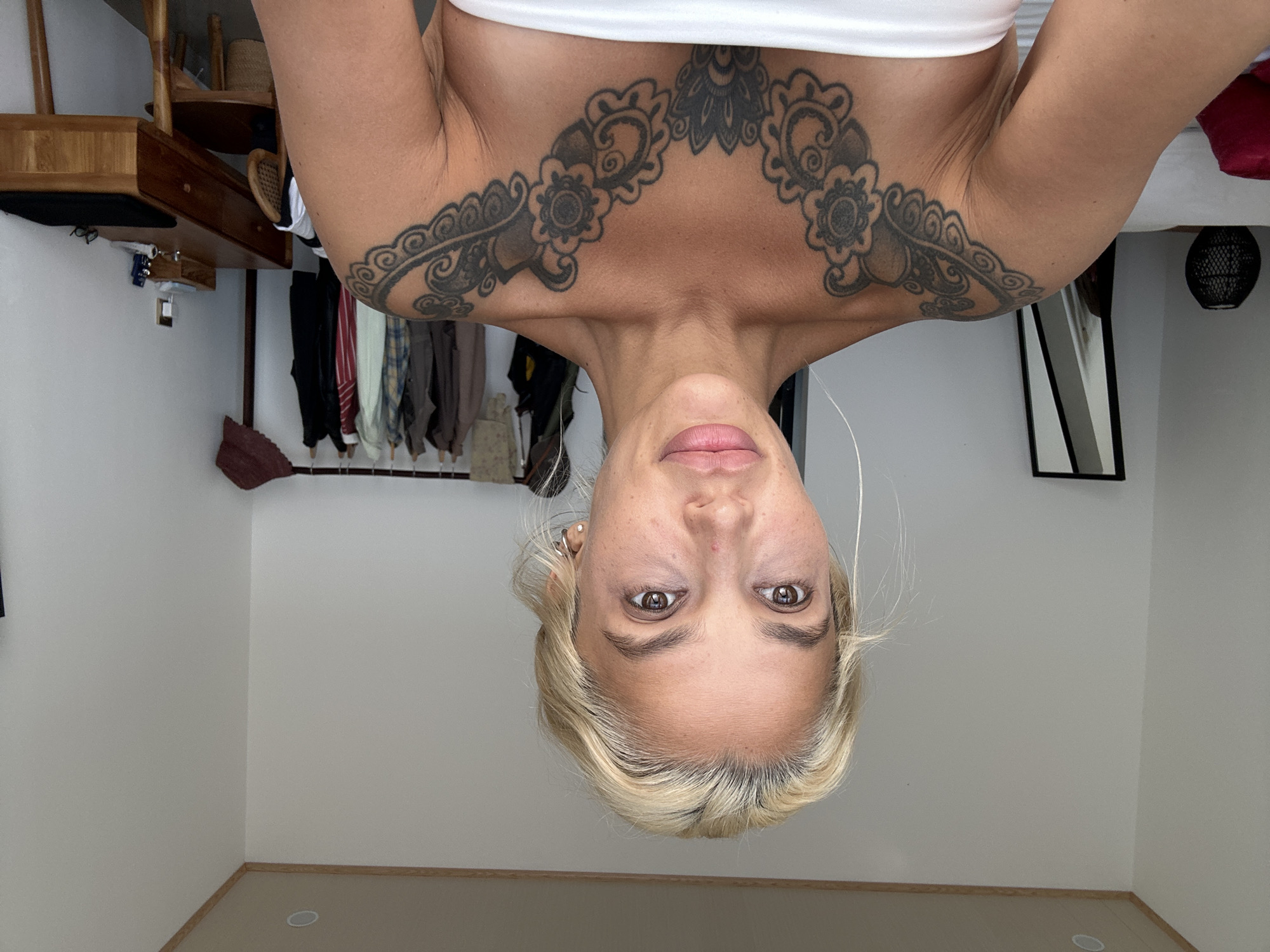 A photograph, oriented with its original top at the bottom, depicts a woman with fair skin and blonde hair. She is wearing a white top and features a prominent black tattoo across her upper chest and shoulders. The background shows a room with light-colored walls. In the upper-left of the frame, a wooden dresser and a clothes rack with various garments hanging are visible. A black frame and a dark-colored spherical object are also present on the right side of the image. The setting is a room within a building, likely located in Kuta Selatan, Indonesia, based on the assumption that the image source is accurate. There are no discernible interactions or specific actions visible in the composition.