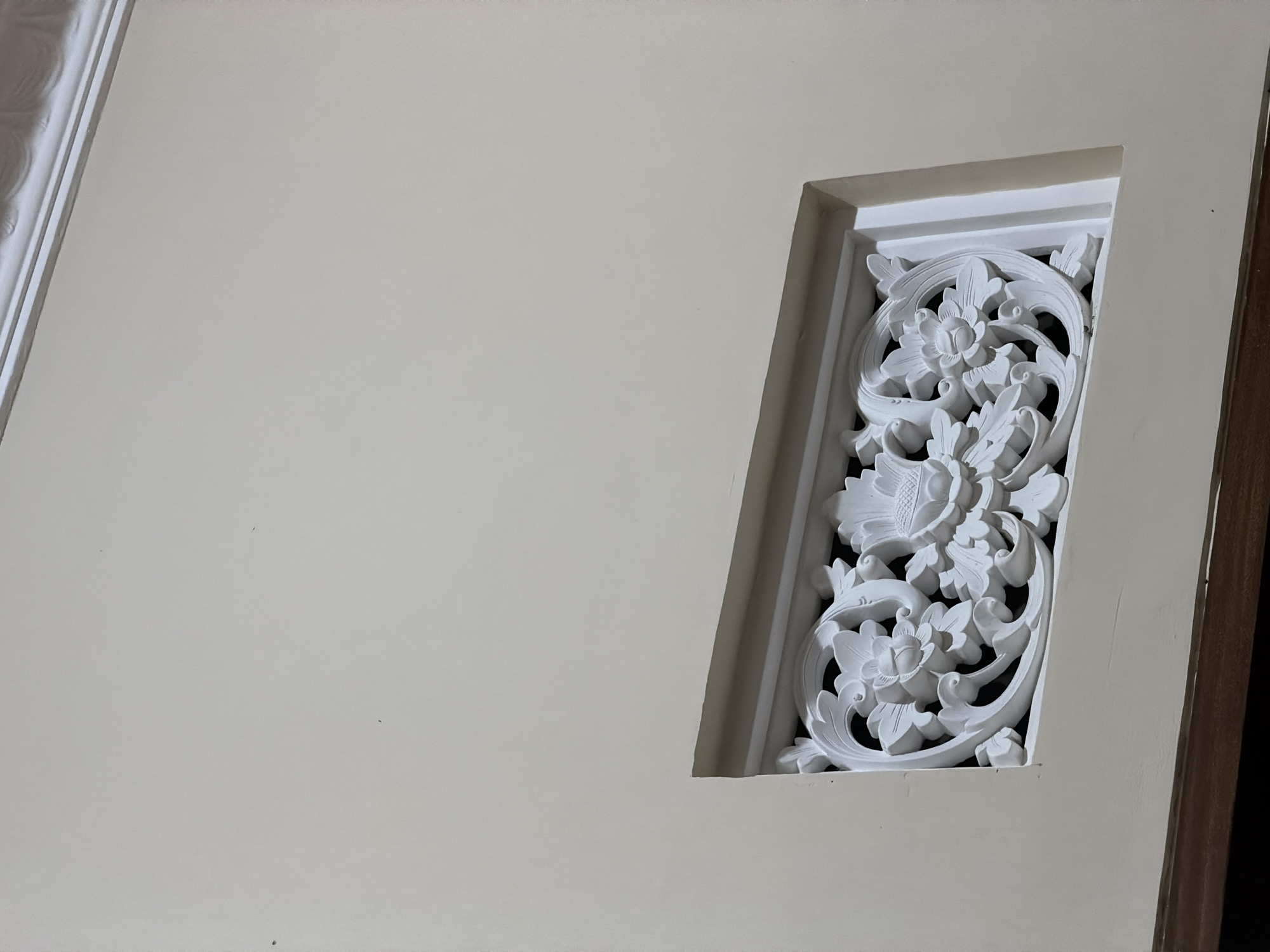 The image shows a close-up, eye-level view of a white ornamental vent cover integrated into a light beige wall. The vent cover features intricate floral and leaf carvings. The wall is smooth and appears to be painted. The top edge of the image shows a portion of white decorative molding. The bottom edge of the image reveals a dark horizontal surface, possibly a wooden trim or furniture. The lighting suggests an indoor setting, casting soft shadows that highlight the relief of the carvings. The image was taken in Wonoayu, Indonesia, and depicts architectural detailing. There are no people or animals visible in the frame.