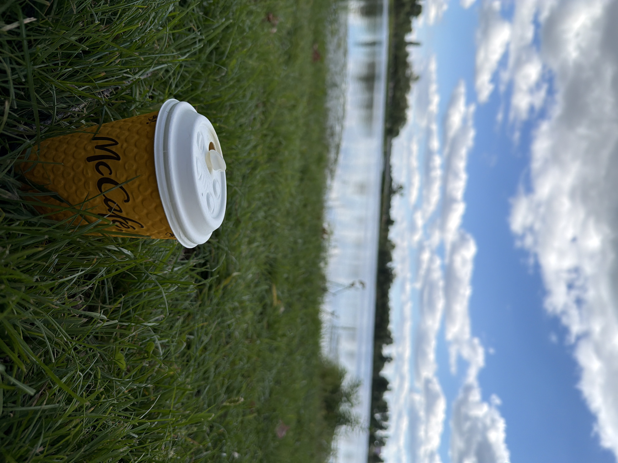 A single McCafe branded disposable coffee cup with a white lid rests on green grass near the edge of a body of water. The cup is yellow with a textured pattern. Beyond the grass, the water reflects the sky, which is filled with scattered white clouds against a blue backdrop. The shoreline features a line of trees. The scene is set in Vyshhorod, Ukraine.