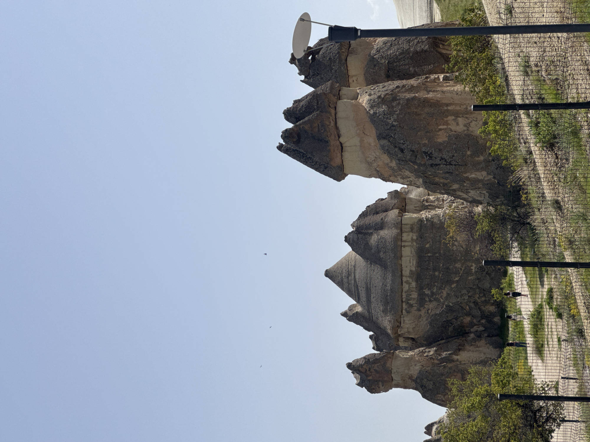 The image captures a unique geological landscape in Avanos, Turkey, featuring the distinctive fairy chimneys. These rock formations, sculpted by erosion, stand against a pale blue sky. A security fence runs along a pathway to the right of the rock formations. A light pole with a reflector rests on one of the chimneys, possibly indicating the area's transformation into a tourist spot.
