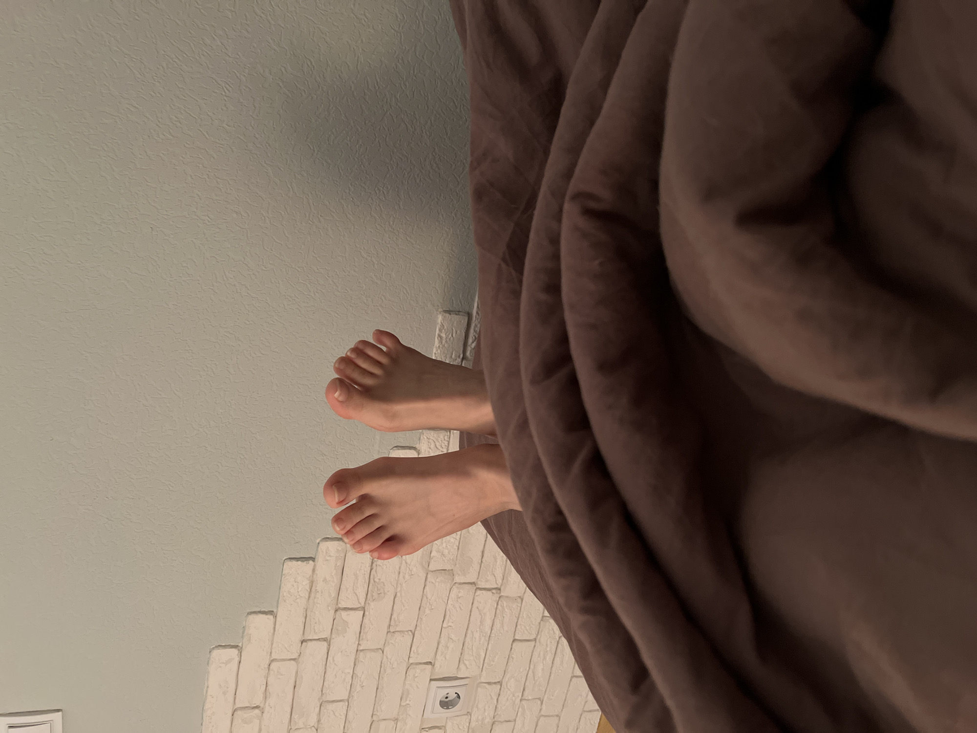 The image shows a pair of bare human feet extending from beneath a brown duvet. The feet are pale, with clearly defined toes. The background includes a light green wall with a textured surface and a white, simulated brick wall. An electrical outlet is visible on the simulated brick wall. The image orientation suggests a view from above, looking down towards the feet. There is no information on location within Saint Petersburg, Russia, included in this image.