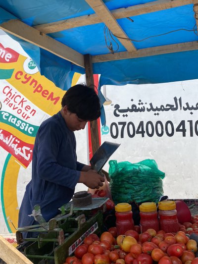 The image shows a vendor at a roadside stall, likely in Kandahar, Afghanistan. The vendor, a young male with dark hair, is wearing a long-sleeved blue shirt and is shown using a large cleaver to cut tomatoes on a dark scale. The stall is covered with a blue plastic tarp and has posters. Visible on the posters are some Arabic writing and the words "Chicken Flavour classic" and "супрем". In front of the vendor, is a pile of tomatoes, along with three jars of red contents with yellow lids. Behind the vendor is a large green plastic bag and another poster with writing. The poster also has the telephone number 0704000410.