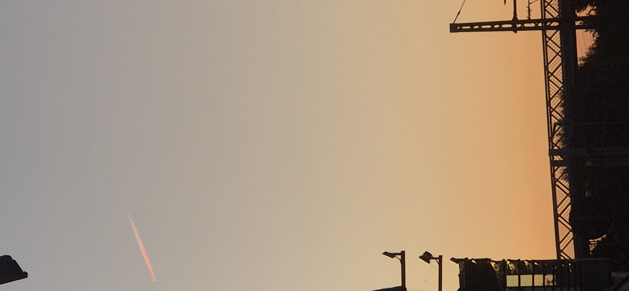 The image shows a sunset with a gradient of colors from pale blue at the top to orange at the bottom. In the upper left portion of the image, a contrail from an aircraft is visible, glowing pink. Silhouetted against the sky are the upper structures of what appear to be railway signals or lighting towers on the left, with a bridge spanning across the middle ground, its lattice structure outlined against the sky. To the far right, another silhouetted industrial structure, possibly a pylon for overhead power lines, is visible. The lower portion of the image is dominated by the dark silhouettes of trees and possibly buildings underneath the bridge. The scene appears to be outdoors, likely in an urban or semi-urban area with transportation infrastructure. While the specific location within Mainz, Germany cannot be determined from the image alone, the presence of railway infrastructure suggests a proximity to train lines or stations.