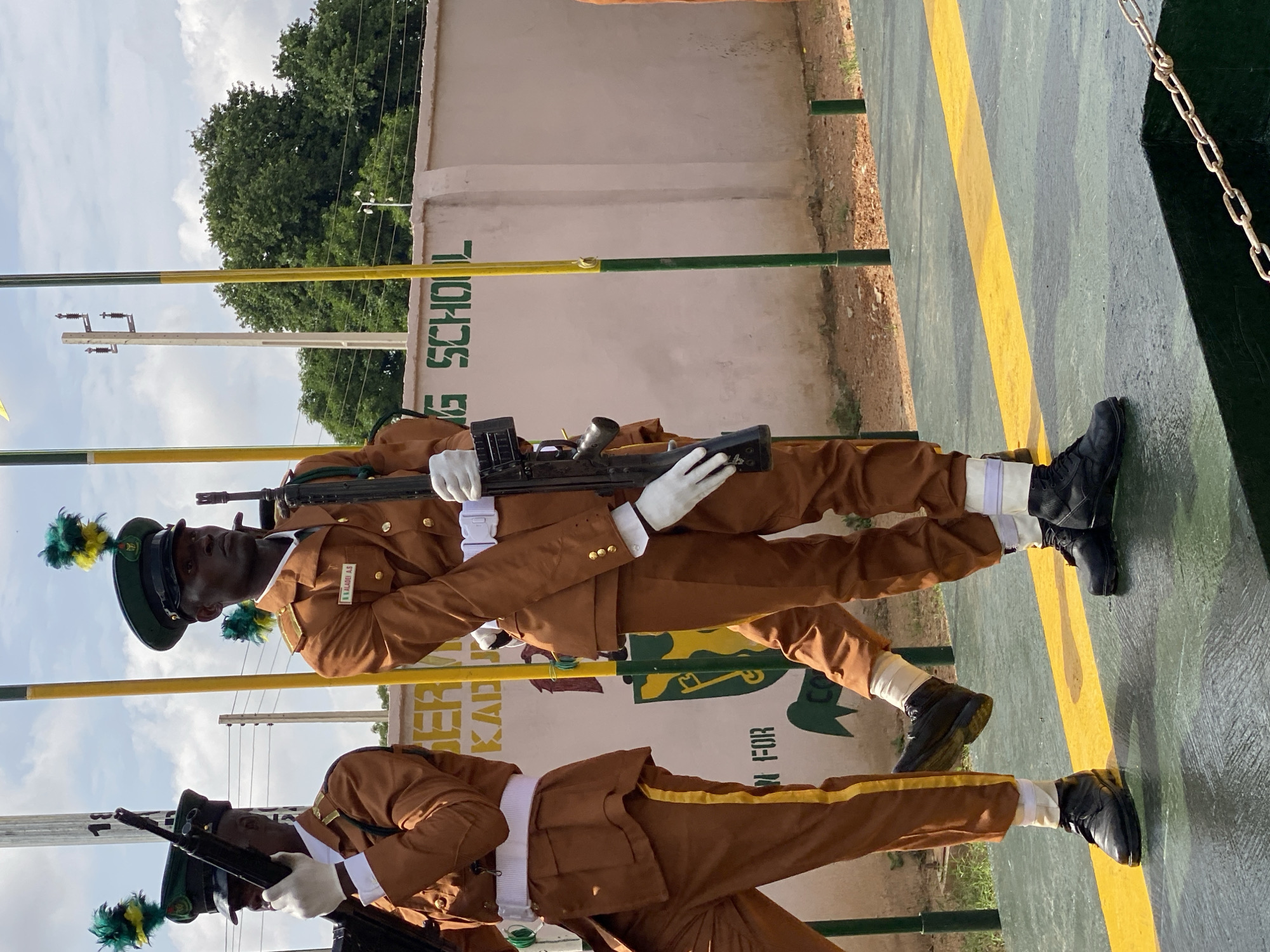 The image shows two people in uniform, likely soldiers or guards, standing at attention outdoors. They are wearing brown uniforms with white belts and white gloves. Both are holding firearms. The person in the foreground is facing left, while the person behind them is facing forward. They are standing on a patterned surface with yellow and green stripes. In the background, there is a light-colored wall with green lettering. Trees are visible behind the wall, and the sky is cloudy. The image appears to have been taken during the day, with diffused light suggesting overcast conditions. The overall scene depicts a formal military or ceremonial occasion. Visible text on the wall reads "NG SCHOOL" and possibly "KADUNA". Another partially visible sign reads "NA ALADEI A.S".