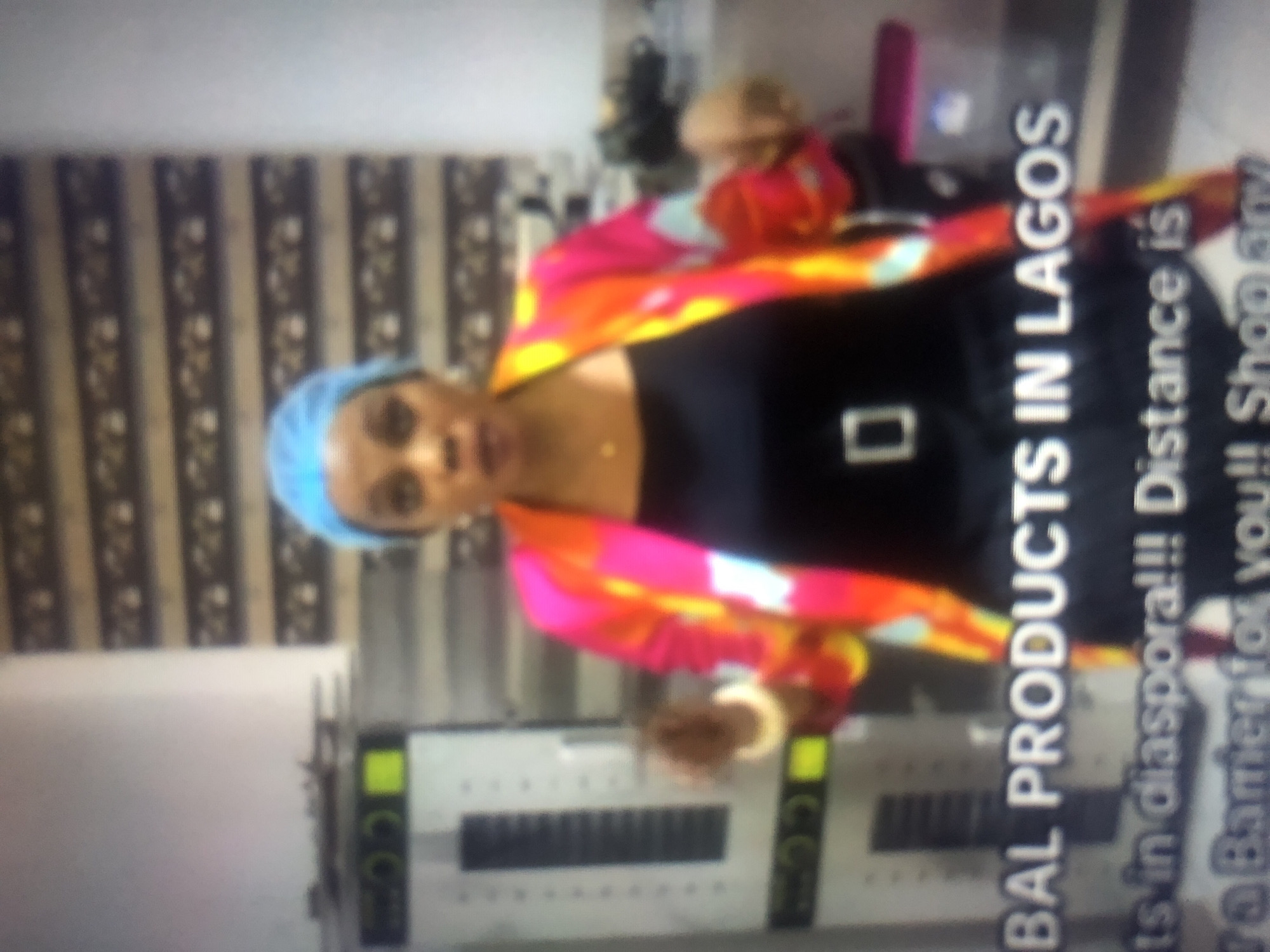 The image shows a woman wearing a blue hairnet and a colorful jacket over a black top, standing in front of industrial equipment. Text on the screen reads "GLOBAL PRODUCTS IN LAGOS" and "its in diaspora!!! Distance is a Barrier for you!! Shop any". The background suggests an indoor setting, possibly a commercial or industrial space, with stacked items and patterned wallpaper. The lighting is somewhat harsh, and the overall image quality is blurred. There is no visible indication of the city of Hawayi, Nigeria.