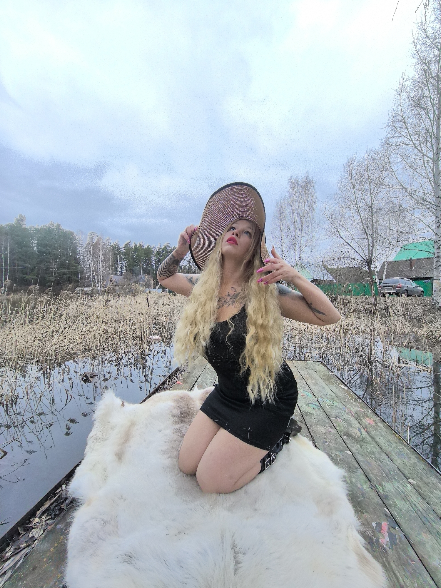 A woman with long, curly blonde hair, wearing a black mini-dress and a large, wide-brimmed straw hat, kneels dramatically on a white fur throw laid over a weathered wooden pier. She has bright red lipstick and long pink fingernails, and visible tattoos adorn her arms and upper chest. Her gaze is directed upwards, and her left hand holds the brim of her hat while her right hand is raised with fingers spread.
The setting is an outdoor, rural landscape in Noviy, Russia, under an overcast sky during daytime. The wooden pier extends over still water, which is densely populated with dry, reedy vegetation. Beyond the water and reeds, a line of bare deciduous trees and evergreen trees forms the backdrop. In the mid-ground, two houses with distinct bright green roofs and a grey car are visible, suggesting a residential area nearby. The bare trees indicate a time of year like late winter or early spring. No visible text is present in the image.