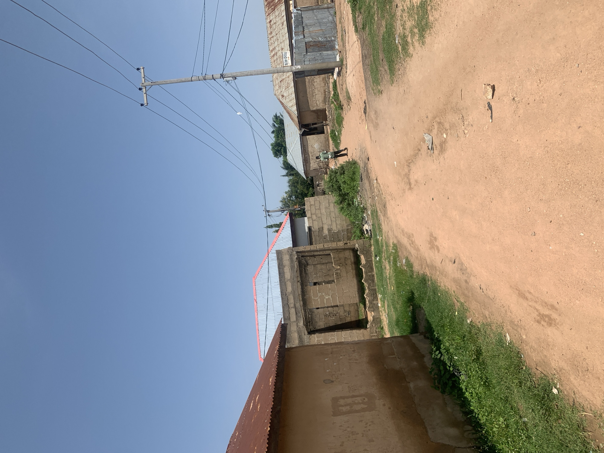 The image depicts a dusty, unpaved road in Jauro Yinu, Nigeria, with buildings on either side. The sky is clear and blue. On the left, a building with a corrugated iron roof and light brown, textured walls is visible. A rectangular window or opening is present on its wall. Further down the road, a partially constructed building made of concrete blocks, with an open doorway, stands. To its right, another building with a light blue corrugated roof with red trim is partially visible. On the right side of the road, several structures with corrugated iron roofs are present, including one with a sign that reads "DESTINY SCHOOLS." A person wearing a green and white checkered shirt and dark pants is walking away from the camera down the road, towards the buildings on the right. A wooden utility pole with multiple power lines stands near the center-right of the image. The ground is predominantly red dirt, with patches of green grass and scattered debris.