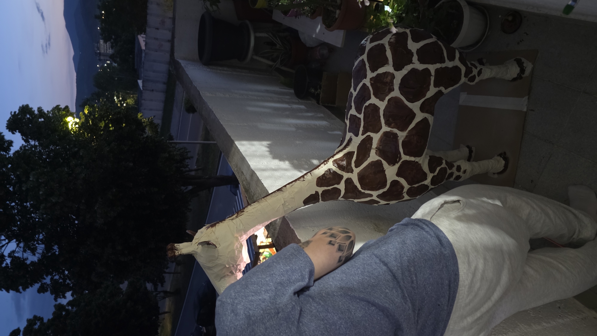 The image depicts a large, giraffe sculpture constructed from papier-mâché placed on a balcony or patio. The sculpture is painted with a giraffe's characteristic brown spots on a cream background. The balcony is narrow and adjacent to a building exterior, and several potted plants are visible along the balcony's edge. A person is visible in the foreground, wearing a gray sweatshirt and light-colored sweatpants. The person is lying down and appears to be interacting with the giraffe's head. The sky is visible in the background along with mountains. The general location is identified to be Blagoevgrad, Bulgaria. Streetlights can be seen through the trees. The time is likely evening or night.