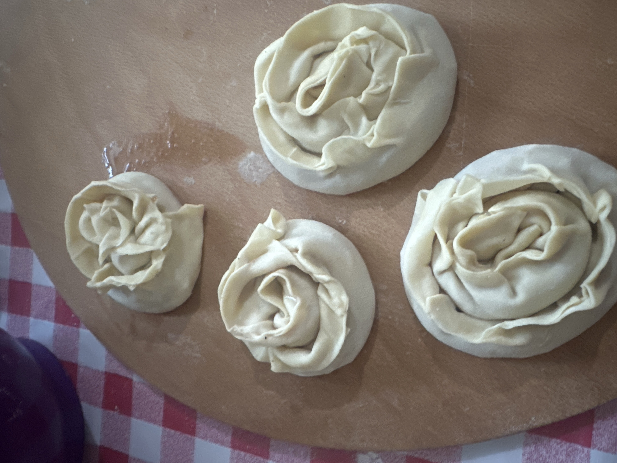Four rose-shaped pastry doughs are arranged on a wooden cutting board. The doughs are light beige in color and appear to be unbaked. They are positioned in a diagonal line from the top left to the bottom right of the frame. In the background, a red and white checkered tablecloth is visible, along with a portion of a dark purple bowl. The scene is set on a wooden surface, likely a kitchen counter or table, in a domestic setting, possibly in Küçükçekmece, Turkey, though no specific location details are discernible from the image.