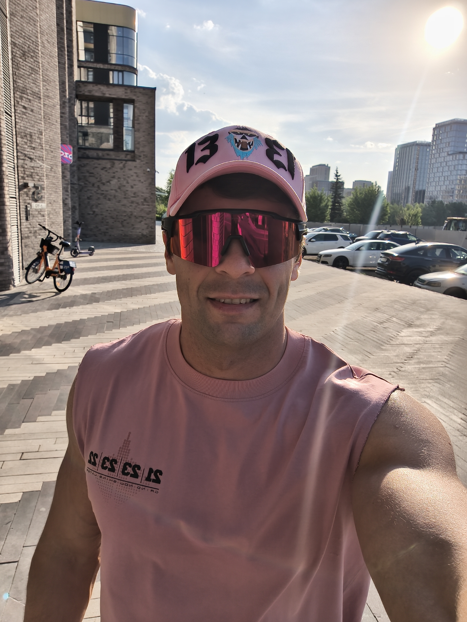 A fair-skinned man is taking a selfie outdoors on a paved area. He is wearing a pink sleeveless shirt, a pink baseball cap with a logo, and pink sunglasses. Behind him is a brick building on the left, a parking lot with cars on the right, and buildings in the background. The buildings and general layout suggest that the location is in the city of Moscow, Russia. In the background, a bicycle and scooter can be observed.