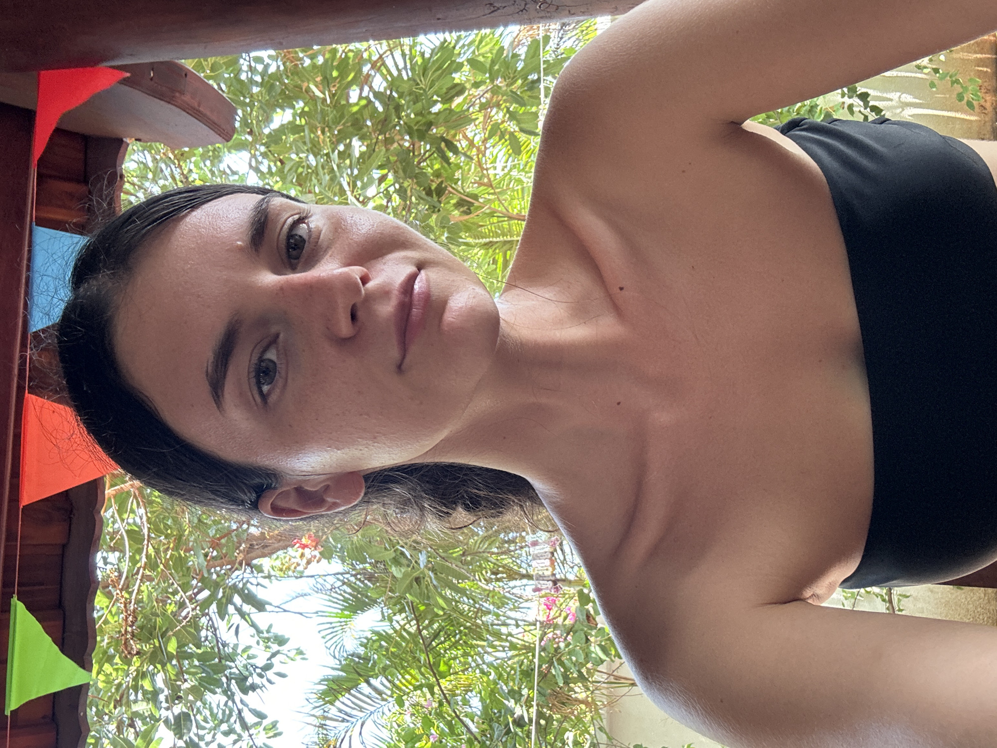 A selfie of a person with dark hair and light skin is taken from a low angle, looking up. The person is wearing a black strapless top. The background features green foliage and a structure decorated with colorful pennant banners. It is difficult to pinpoint the precise location within Mũi Né based on the image alone. However, the presence of lush vegetation suggests a location near a garden, resort, or natural area.