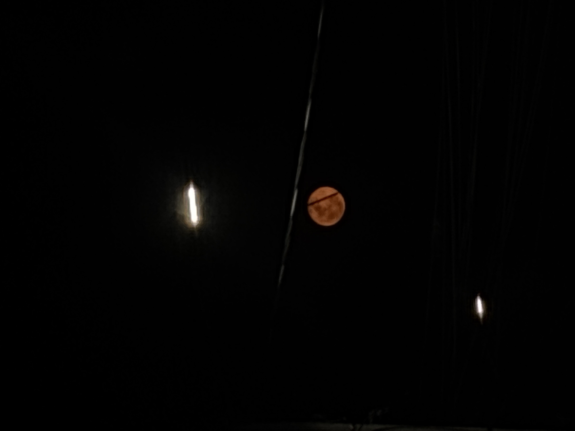 The image, taken at night, shows a full, orange moon in the sky. Several power lines crisscross the foreground. To the left of the moon, a bright light source is visible, likely a street light. Another, dimmer light source appears on the right side of the image. The background is a dark, night sky. The location cannot be determined from the information provided.