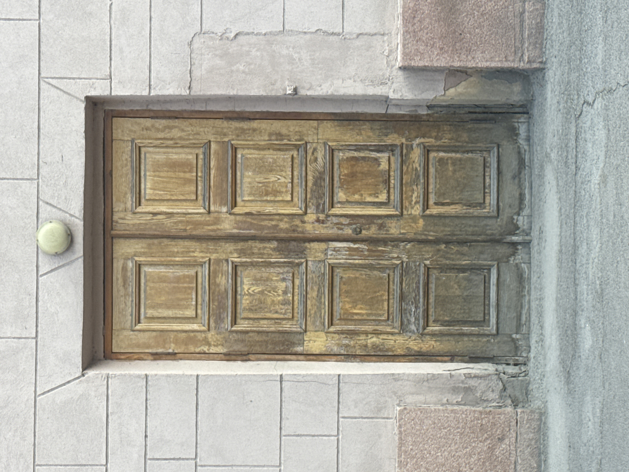 This is a medium shot, eye-level, outdoor, vertical, eye-level, front view, daylight photograph of a weathered wooden double door set into a light-colored stone brick wall. The door is divided into six rectangular panels, with the top three panels being smaller than the bottom three. The wood has a visible grain and appears to be aged, with some areas showing wear and tear. Above the door, centered, is a round, light-colored fixture, possibly a light or an intercom. The stone brick wall is textured and appears to be made of large, rectangular blocks. To the left and right of the door, at the bottom, are sections of pinkish-red speckled stone. A grey, textured pavement is visible in the foreground, with a faint crack running across it. The environment suggests an exterior urban setting.